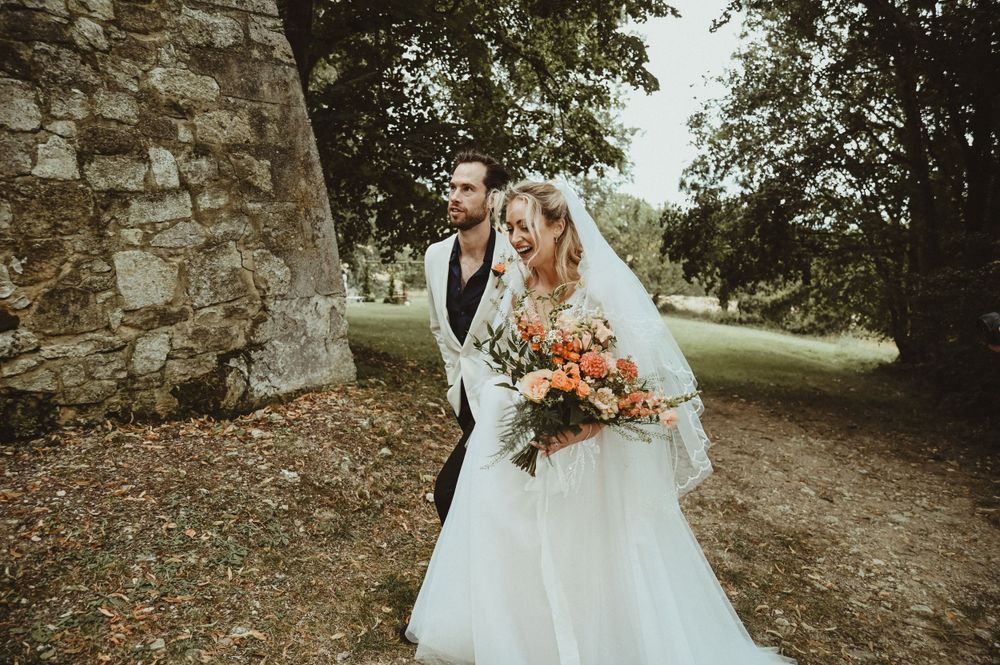 A wedding couple poses near a historic stone wall with autumn-colored bouquet.