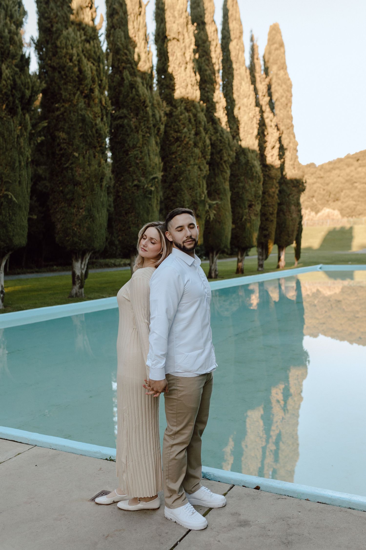 A couple poses beside a tranquil swimming pool surrounded by tall cypress trees at sunset.