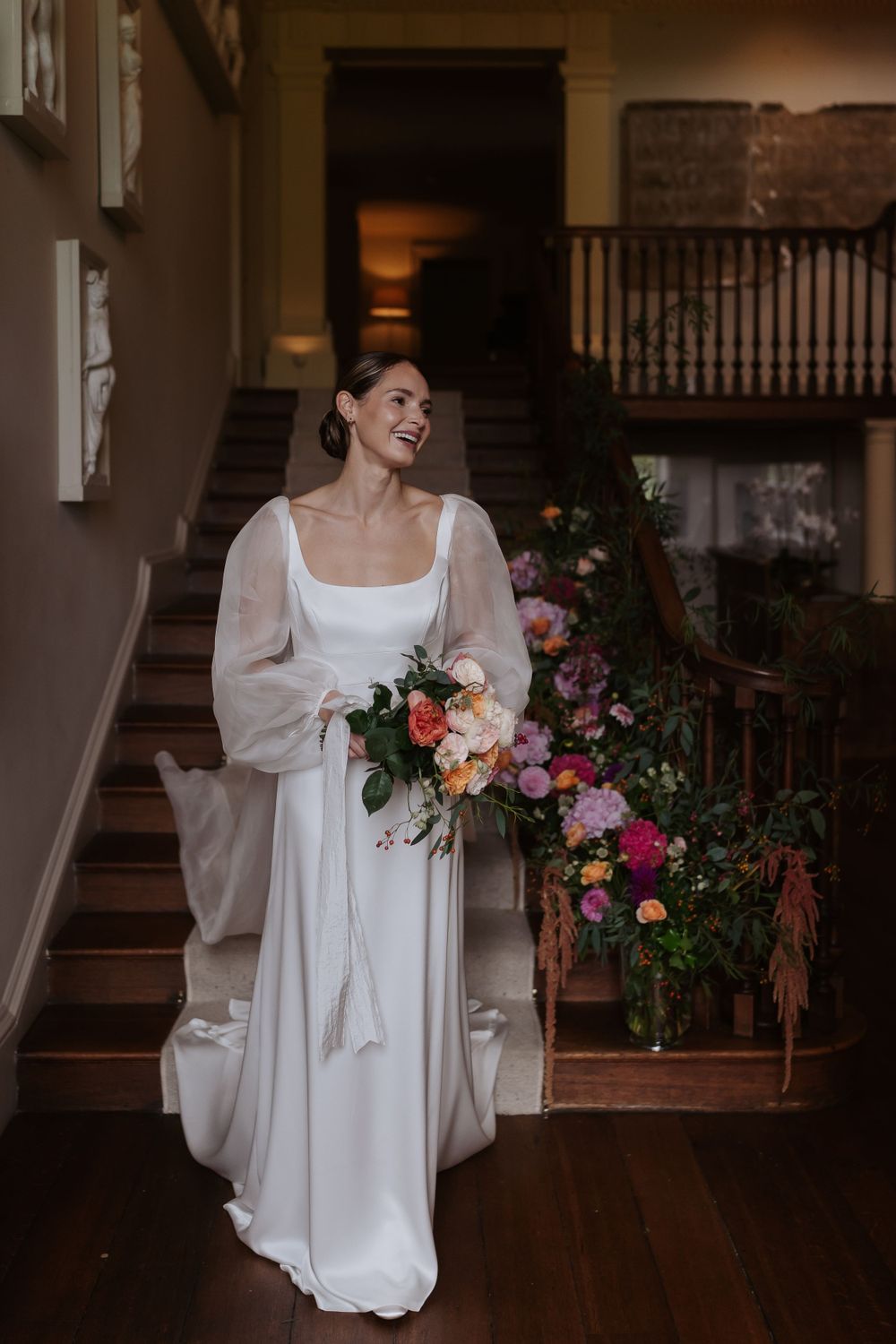 Elegant bride in long-sleeved white dress holds vibrant floral bouquet while standing on wooden staircase with floral decor.