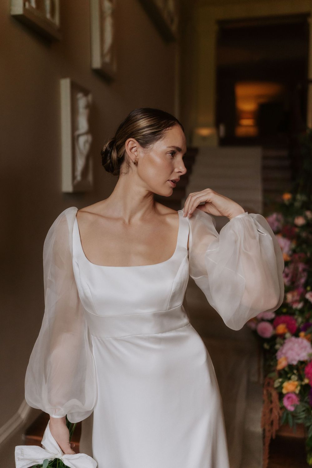 Elegant bride wearing a classic white wedding dress with sheer billowing sleeves poses gracefully against a floral backdrop.