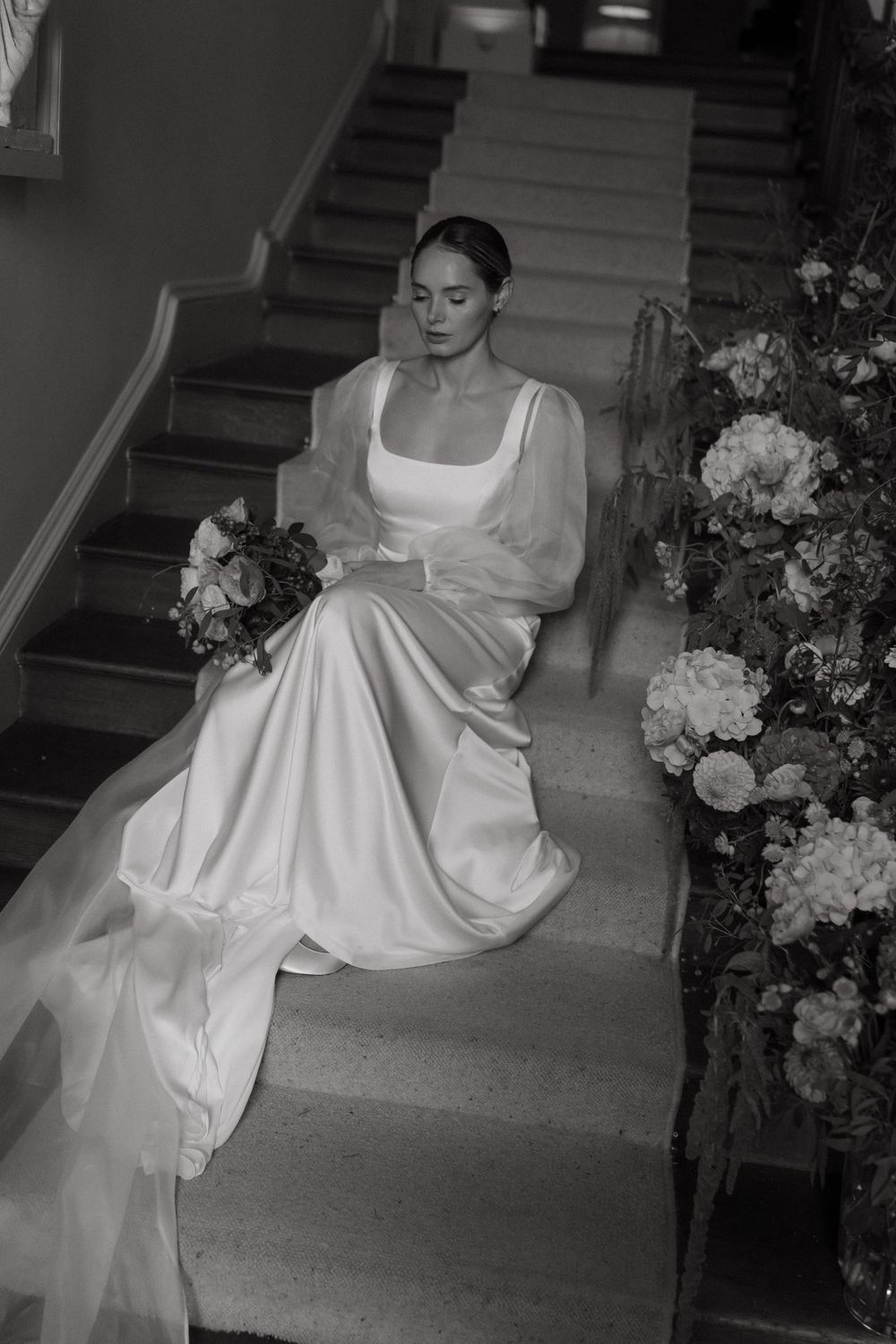 Black and white photo of a bride in an elegant satin gown with sheer sleeves sitting on stairs adorned with floral arrangements.