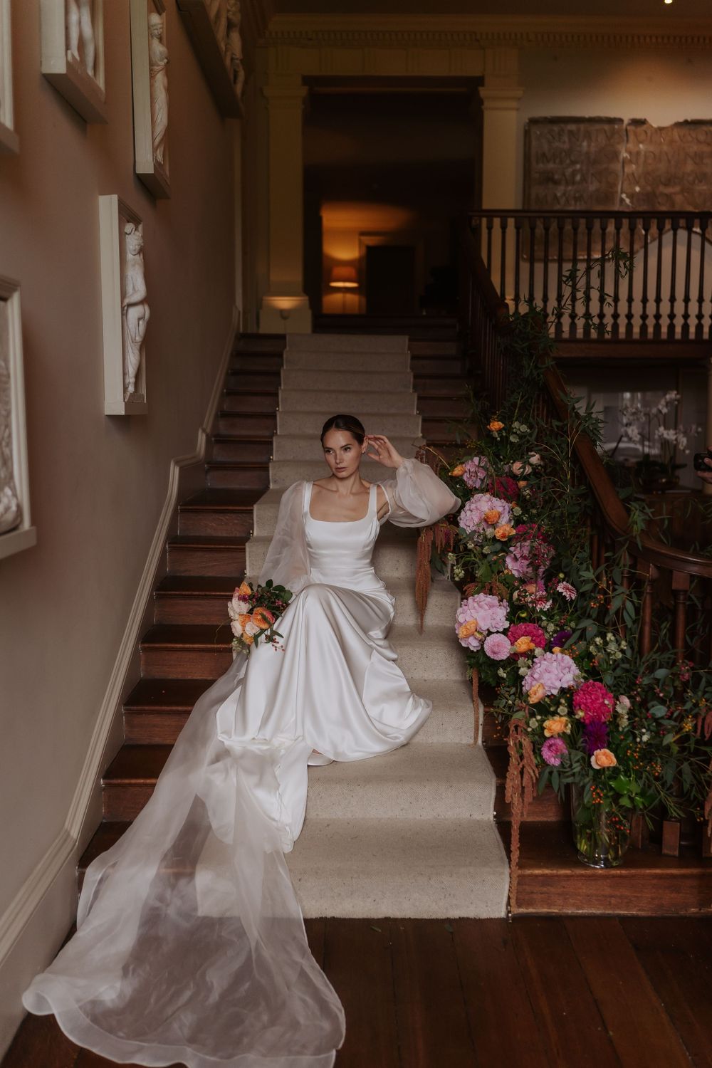 Elegant bride in white satin gown with puff sleeves poses on grand staircase adorned with colorful floral arrangements.
