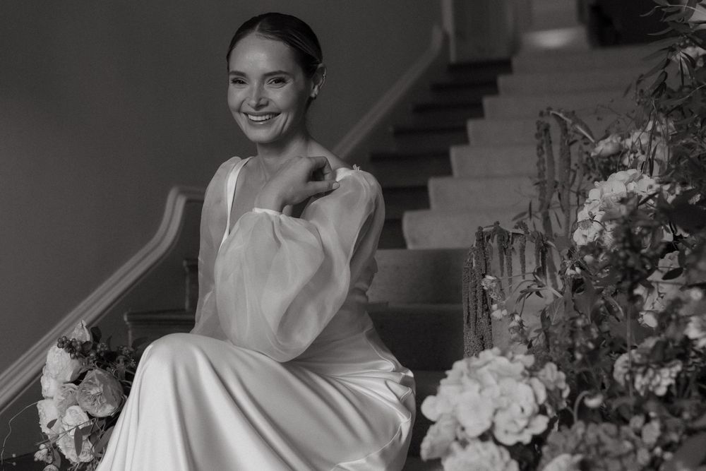 Black and white portrait of bride in elegant white gown seated on stairs near floral arrangements.