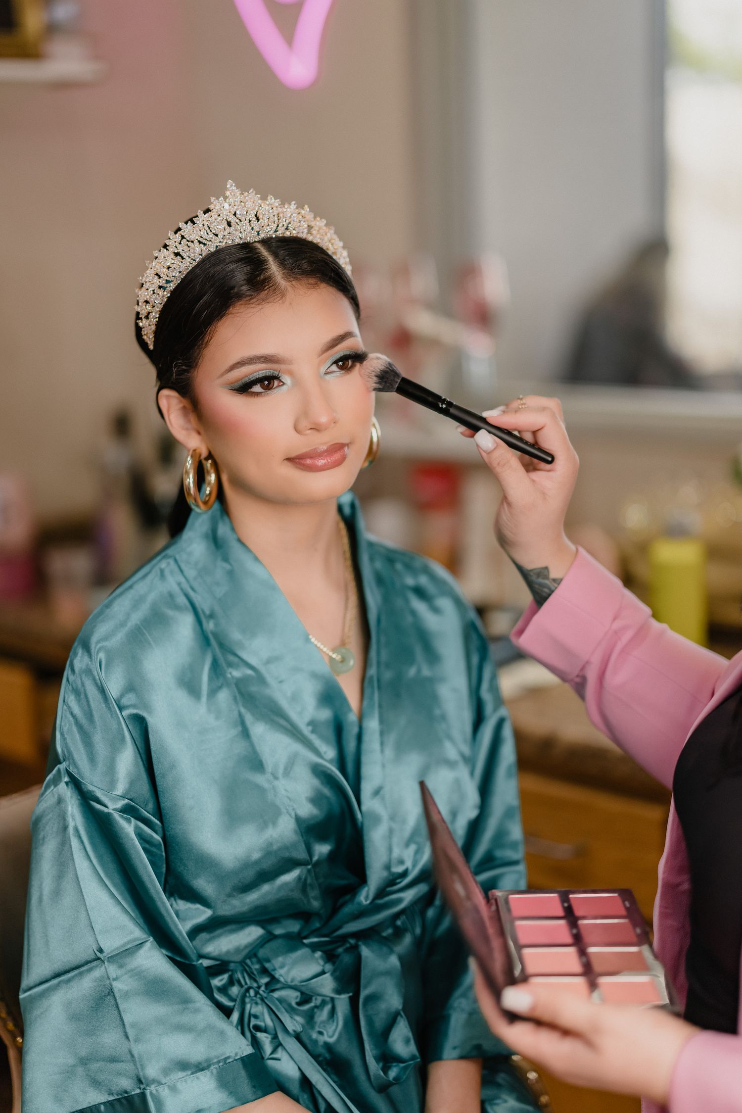 A makeup artist applies cosmetics to someone wearing a teal silk robe and crystal tiara during a beauty session.