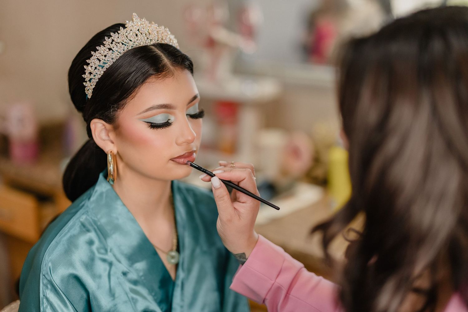 Makeup artist applies lipstick to bride wearing teal silk robe and crystal tiara during wedding preparation.