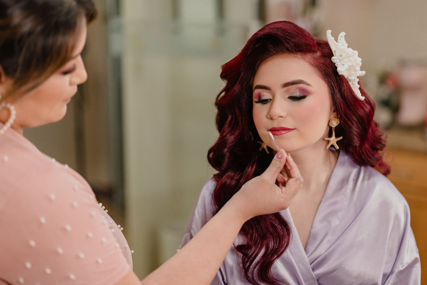 Wedding makeup artist applies finishing touches to model wearing white silk robe with red curled hair and white flower accessory.