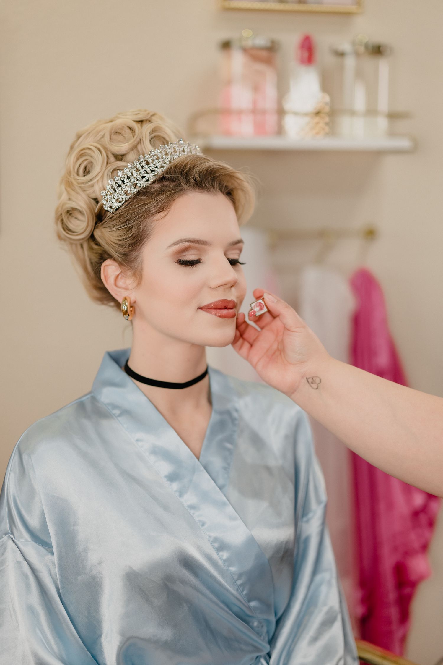 Makeup artist applying finishing touches to elegant bridal look with tiara and blue silk robe.
