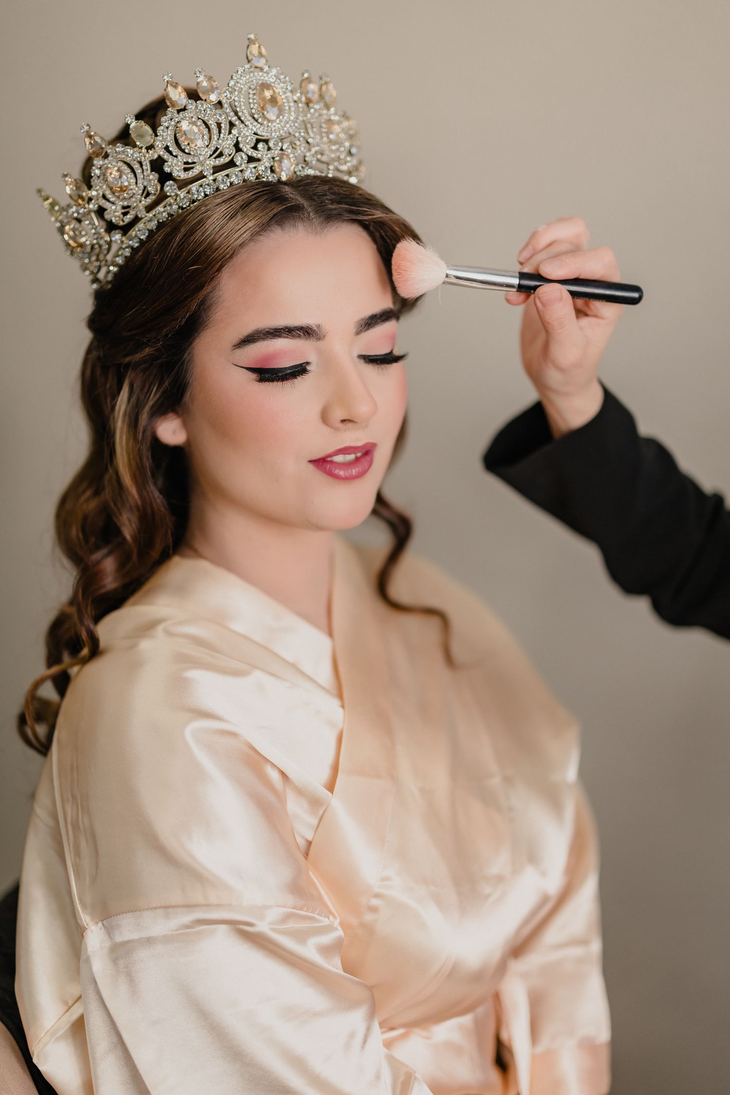 Makeup artist applies eye shadow to model wearing ornate crystal crown and silk robe during bridal preparation.