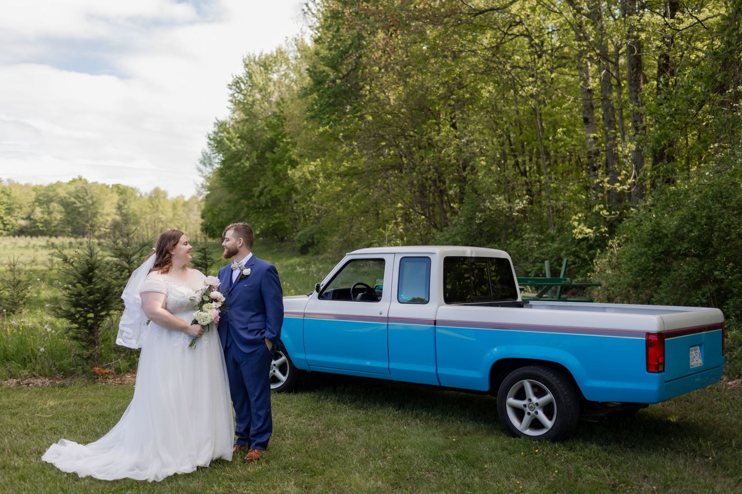 A couple in wedding attire stands in front of a vintage blue and white pickup truck parked on grass near trees.