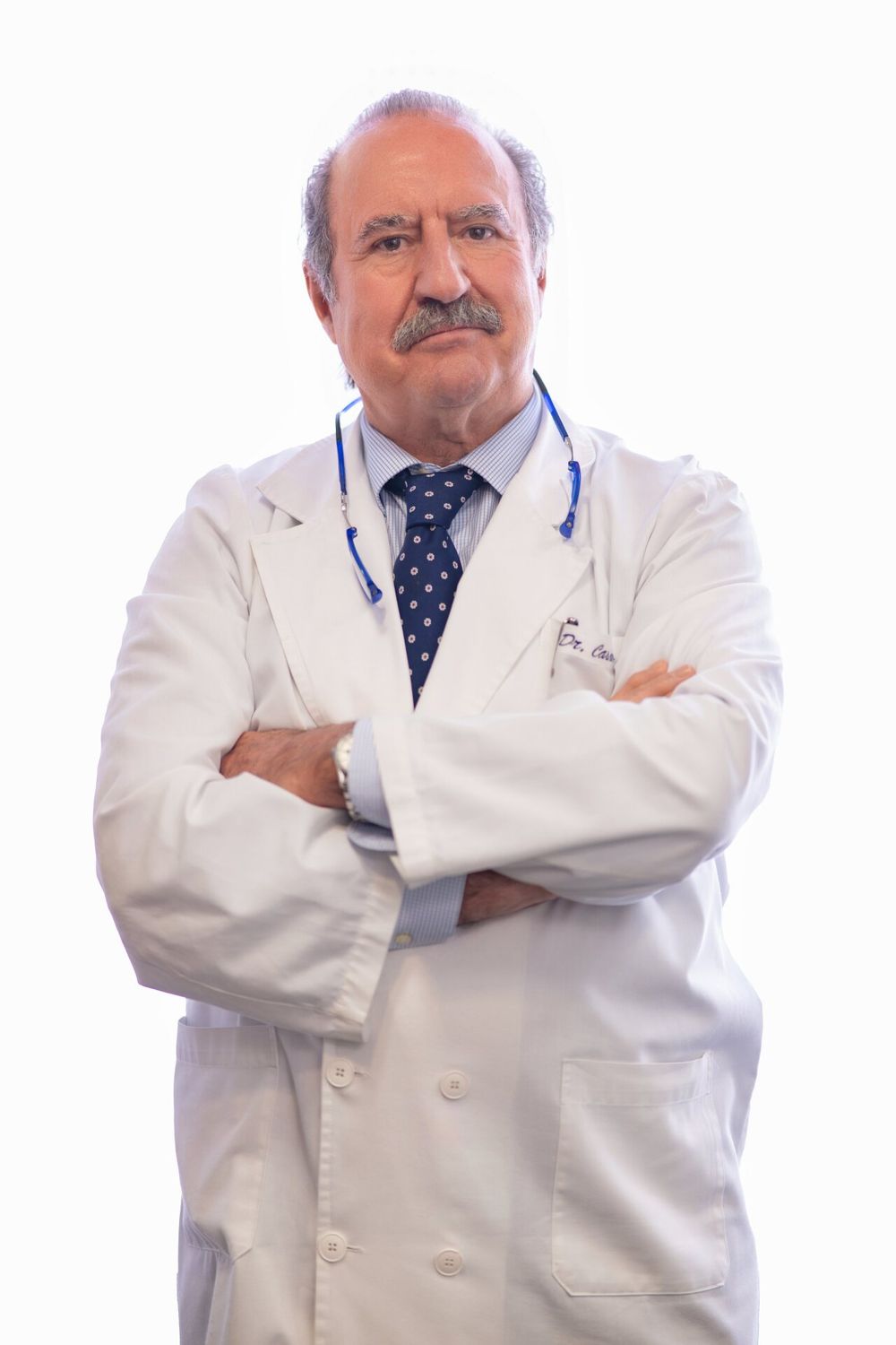 Healthcare professional in white coat and polka dot tie standing with arms crossed in medical setting.