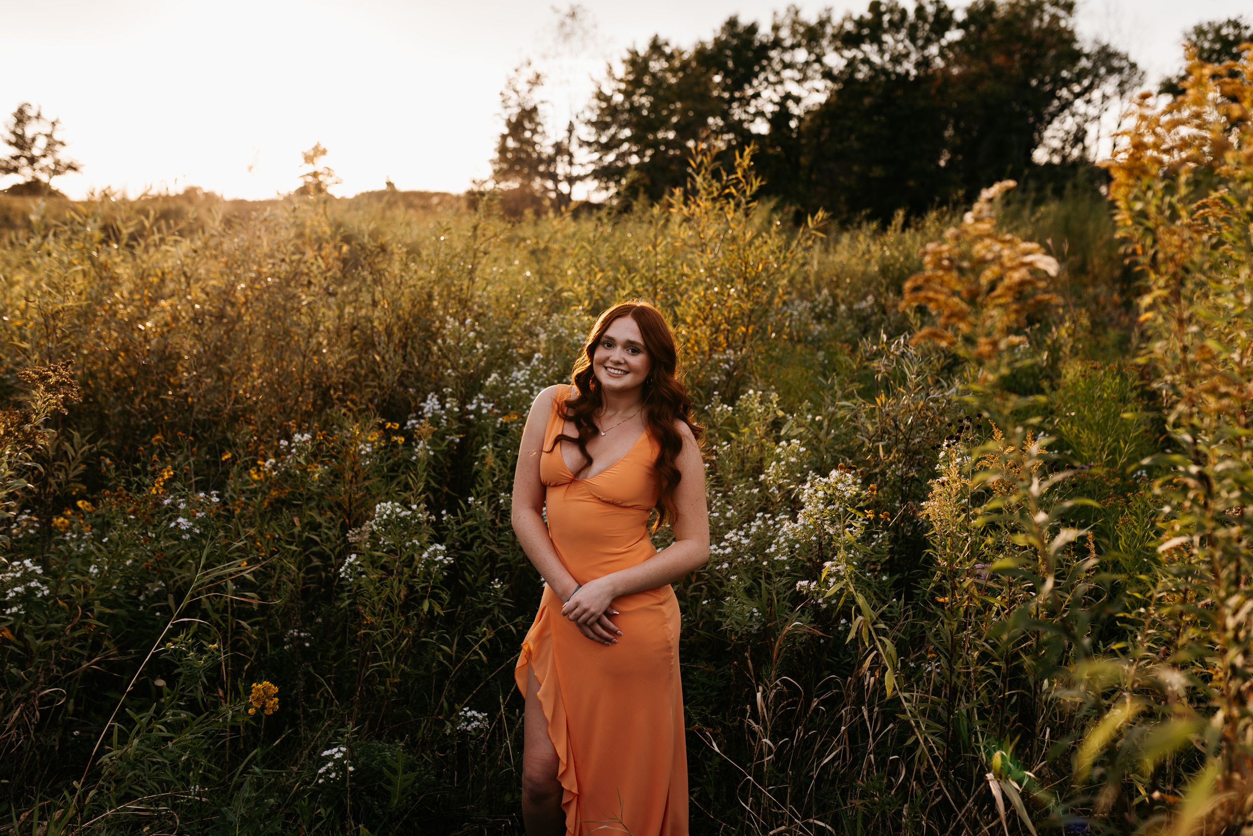 A figure in a rust-colored dress poses in a golden meadow at sunset among tall wildflowers and grass.