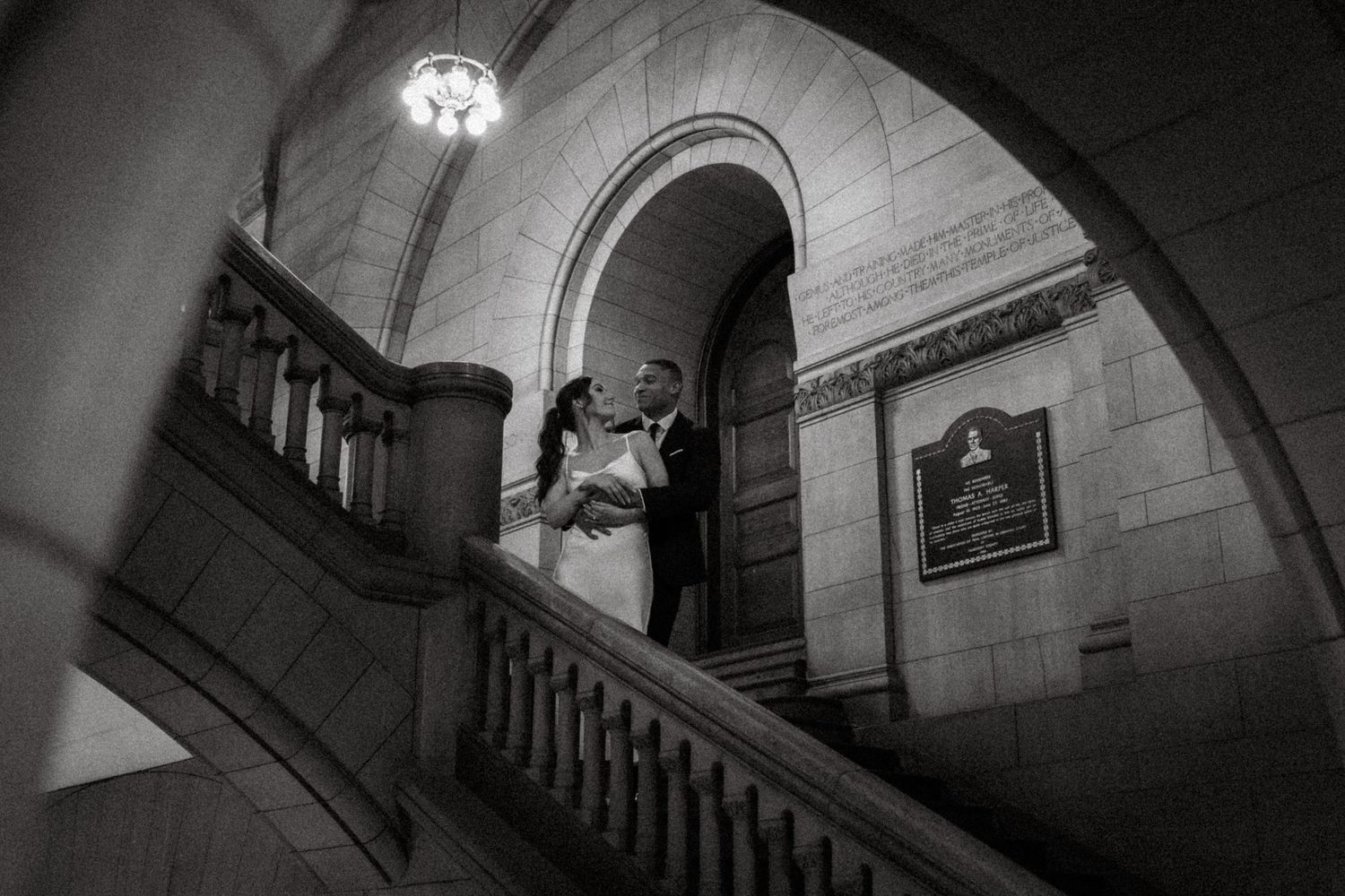 A romantic moment captured in black and white on an elegant stone staircase in a historic building interior.
