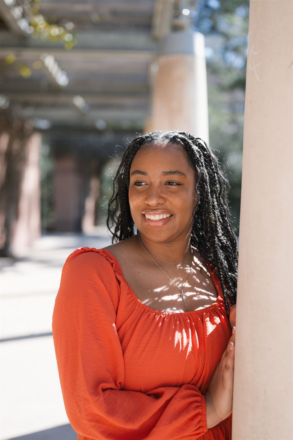 Person in an orange long-sleeved top smiles while standing near a column on a sunny day.