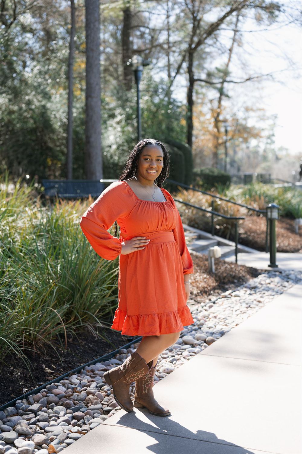 Woman in orange flowy dress with brown boots poses on outdoor path surrounded by trees and rocks.