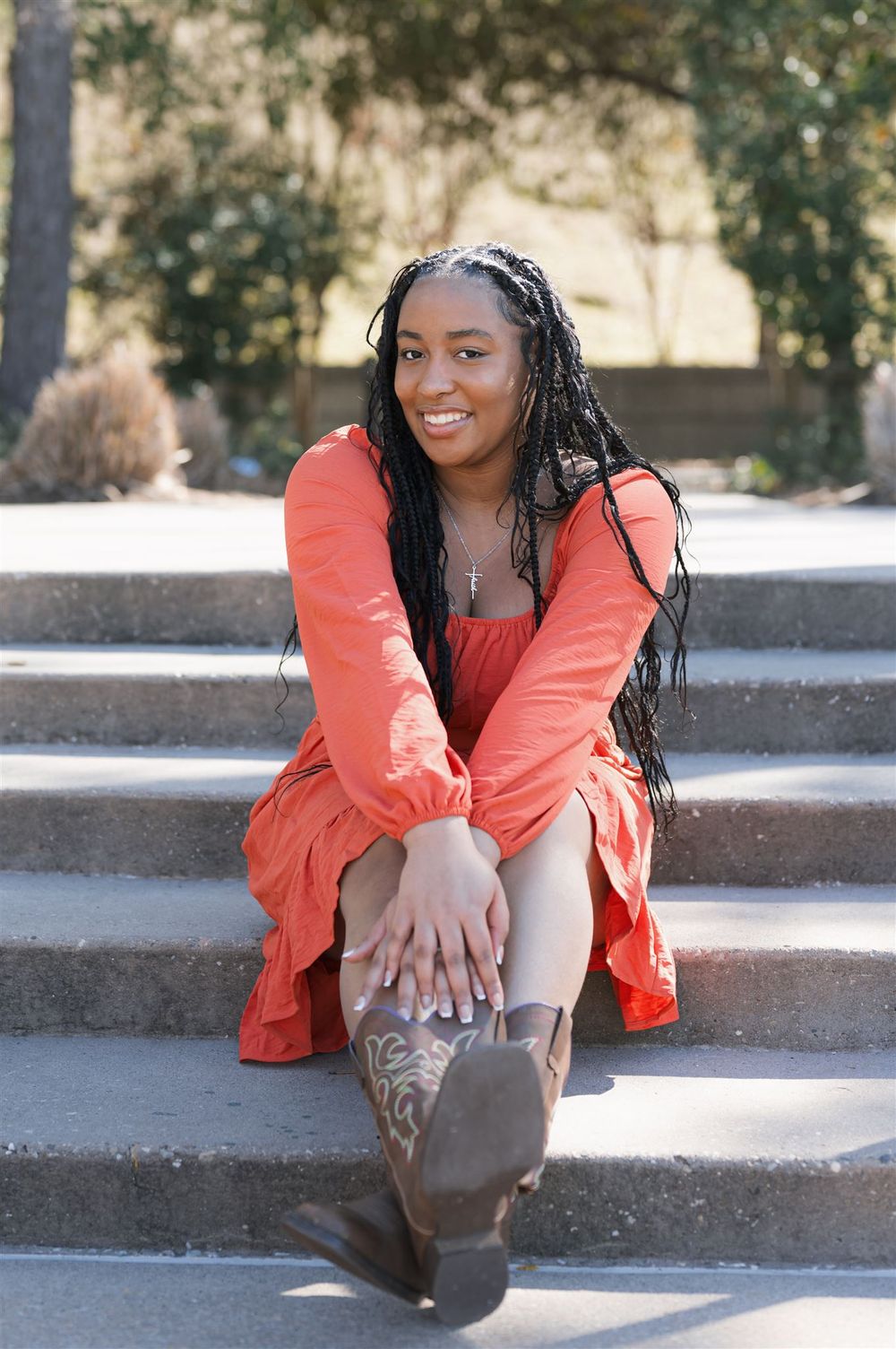 Smiling person sitting on outdoor stone steps wearing a coral-colored top and boots in natural light.