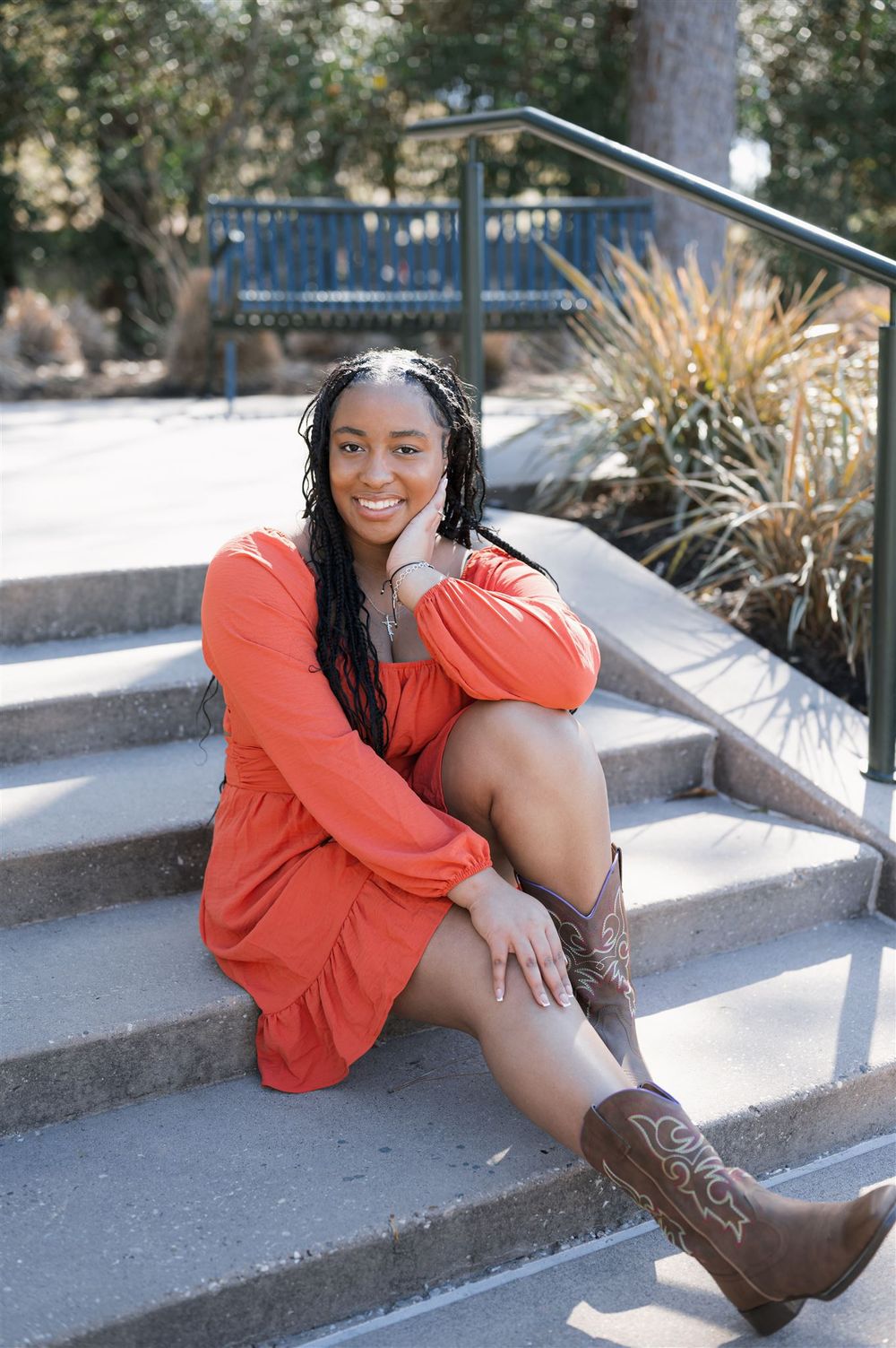 Someone in an orange dress and brown boots sits casually on outdoor concrete steps near ornamental grasses in a sunny setting.