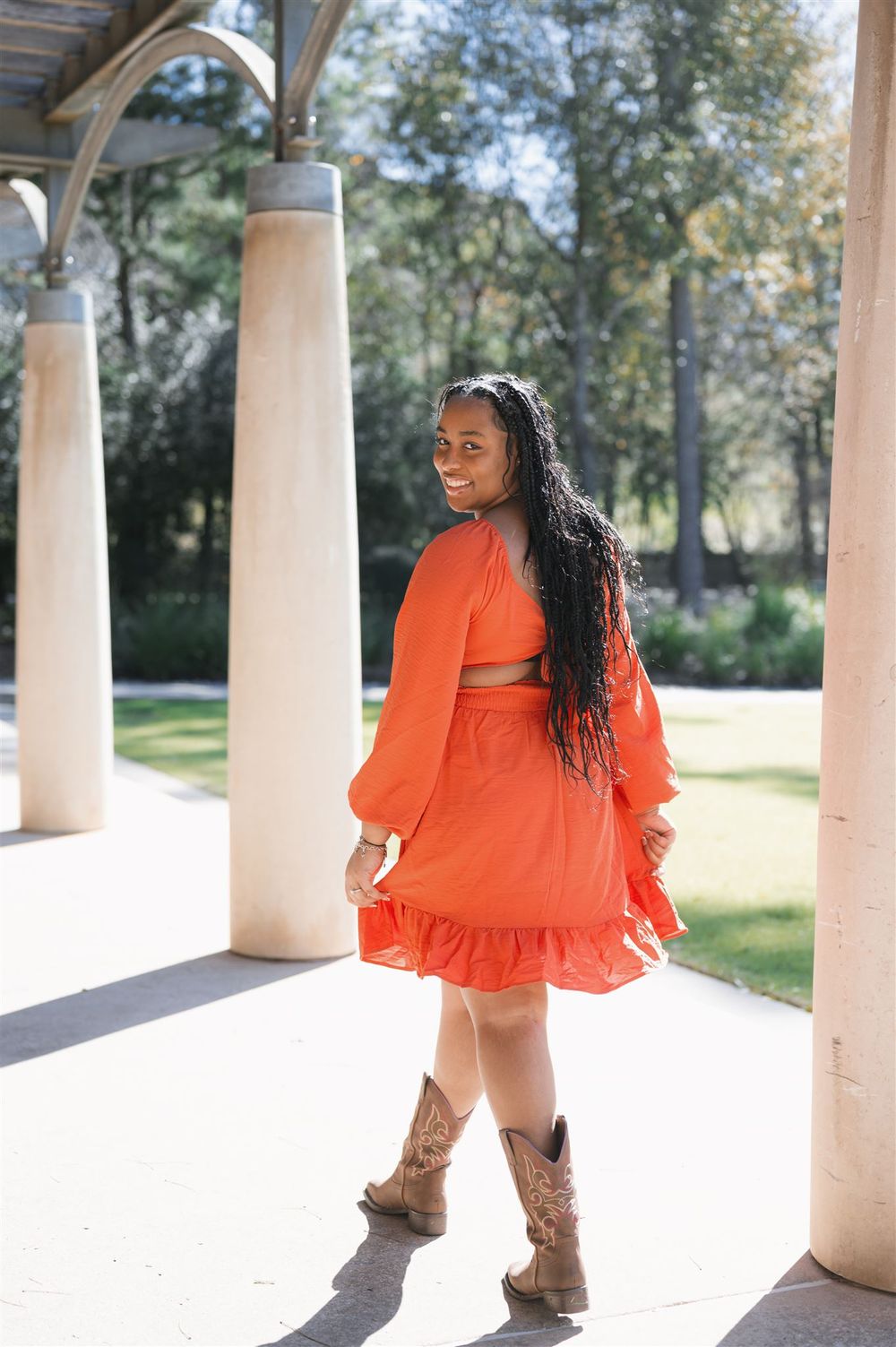A bright orange ruffled dress paired with brown cowboy boots is worn while posing between white columns on a sunny day.