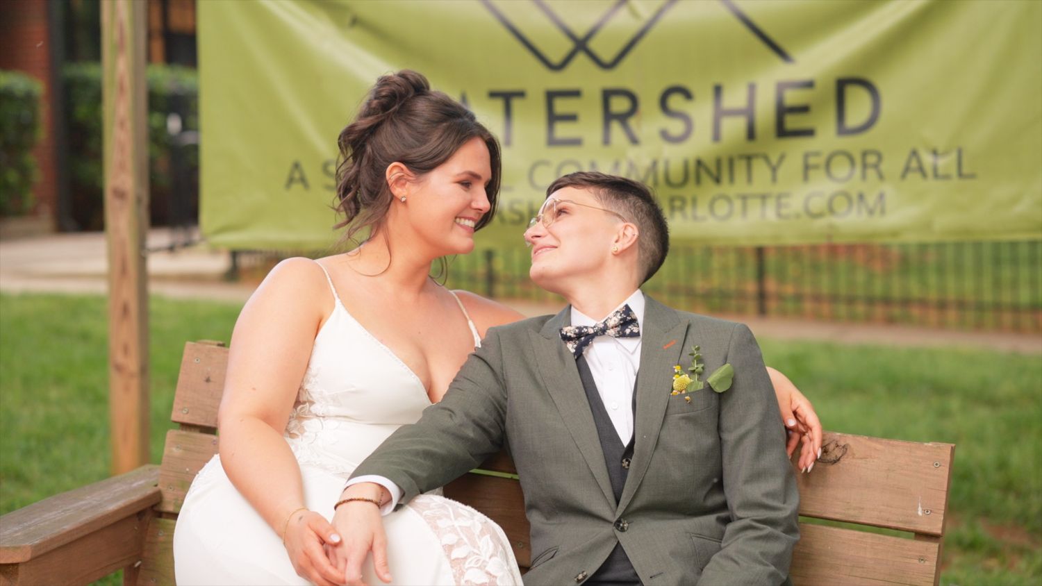 A couple sits together on a wooden bench, sharing a tender moment at an outdoor wedding venue with a banner behind them.