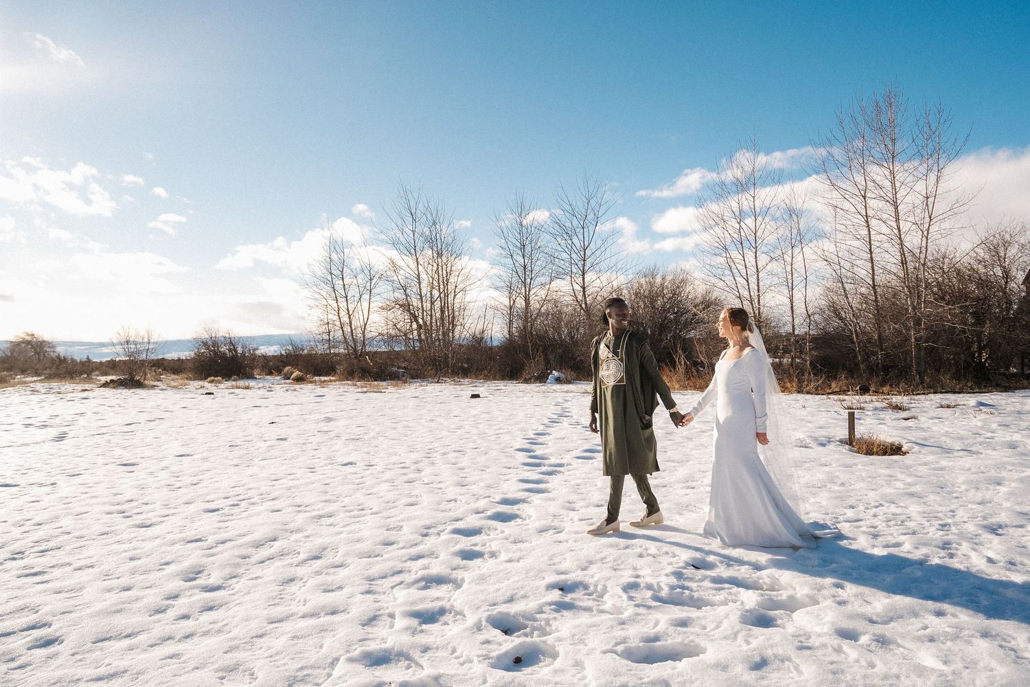 Two people holding hands in a white snowy landscape with bare trees and blue sky on a winter day.