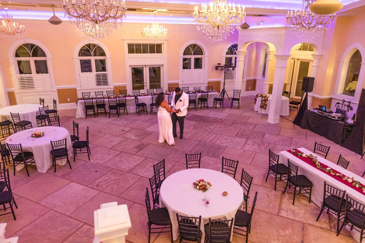 A couple shares a romantic first dance in an elegant ballroom bathed in pink lighting with empty chairs surrounding them.