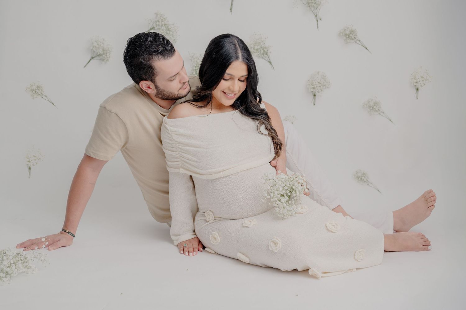 A couple wearing beige clothing poses intimately on a white floor with delicate floral wall decorations.