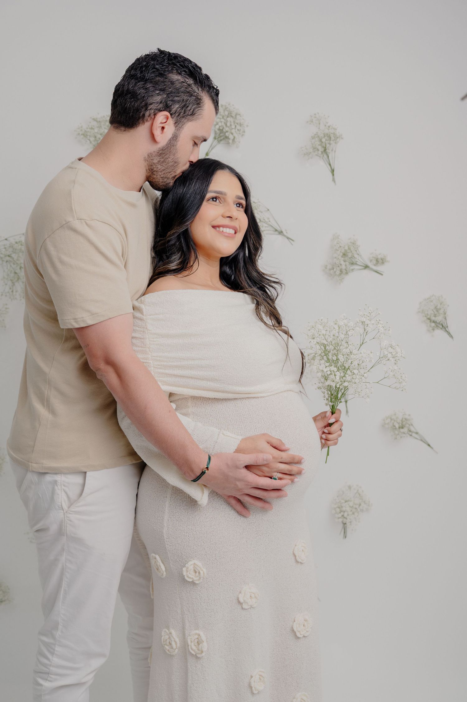 A romantic maternity photo shoot with neutral tones features a couple against a white wall decorated with delicate florals.
