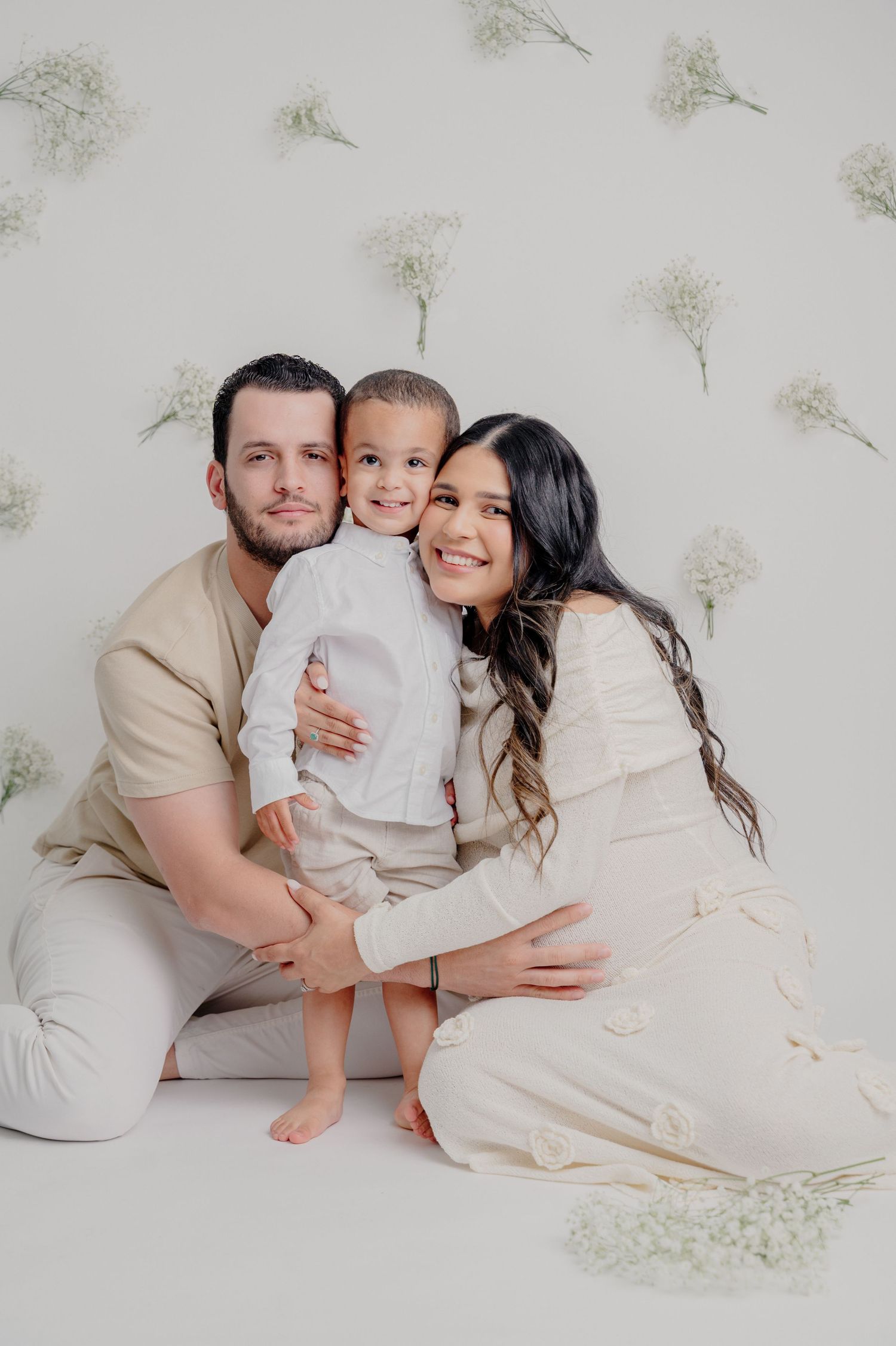 A family poses together wearing neutral tones against a floral wall backdrop in a minimalist studio setting.