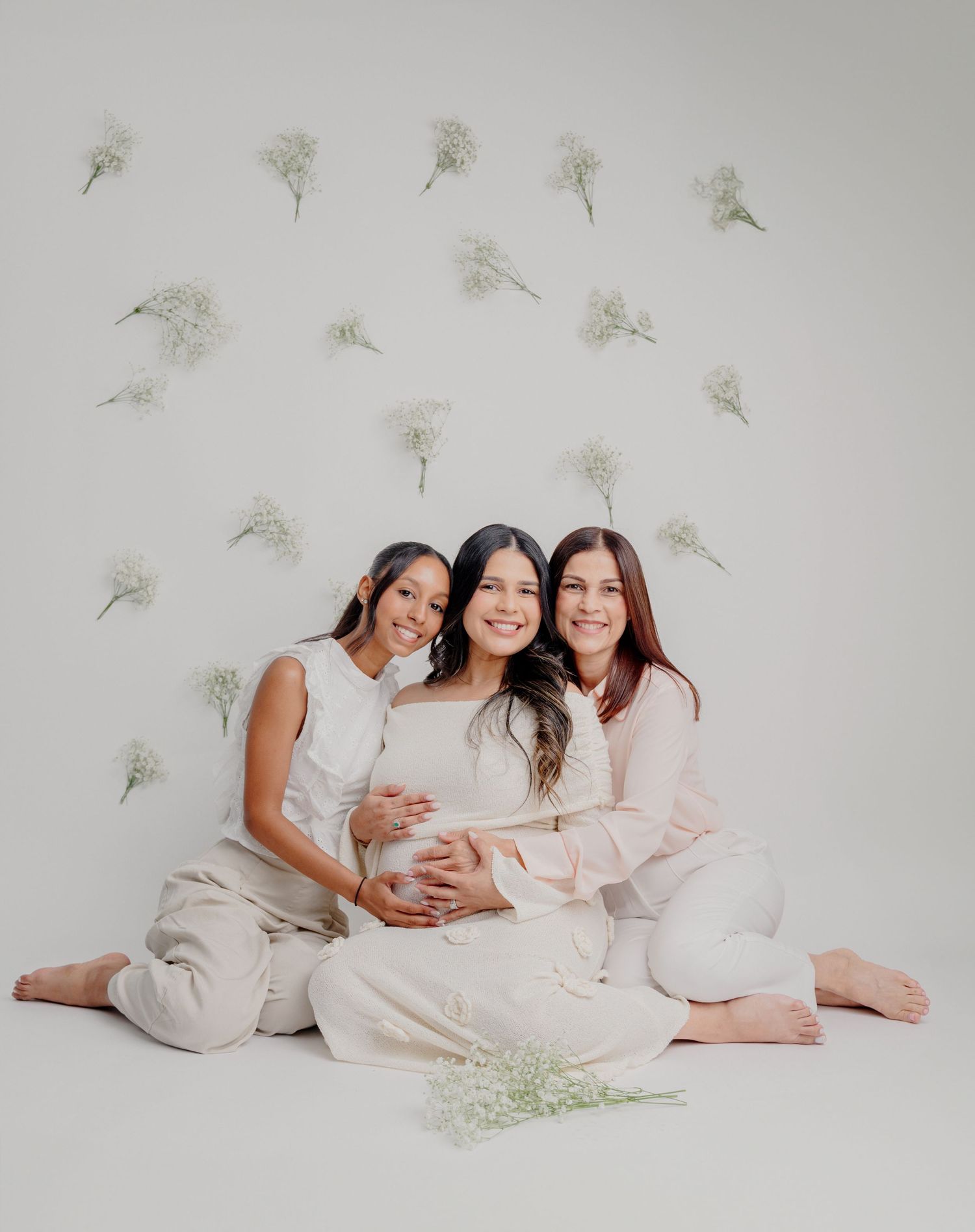 Three women in light colored clothing sit together against a wall with delicate floral decor.