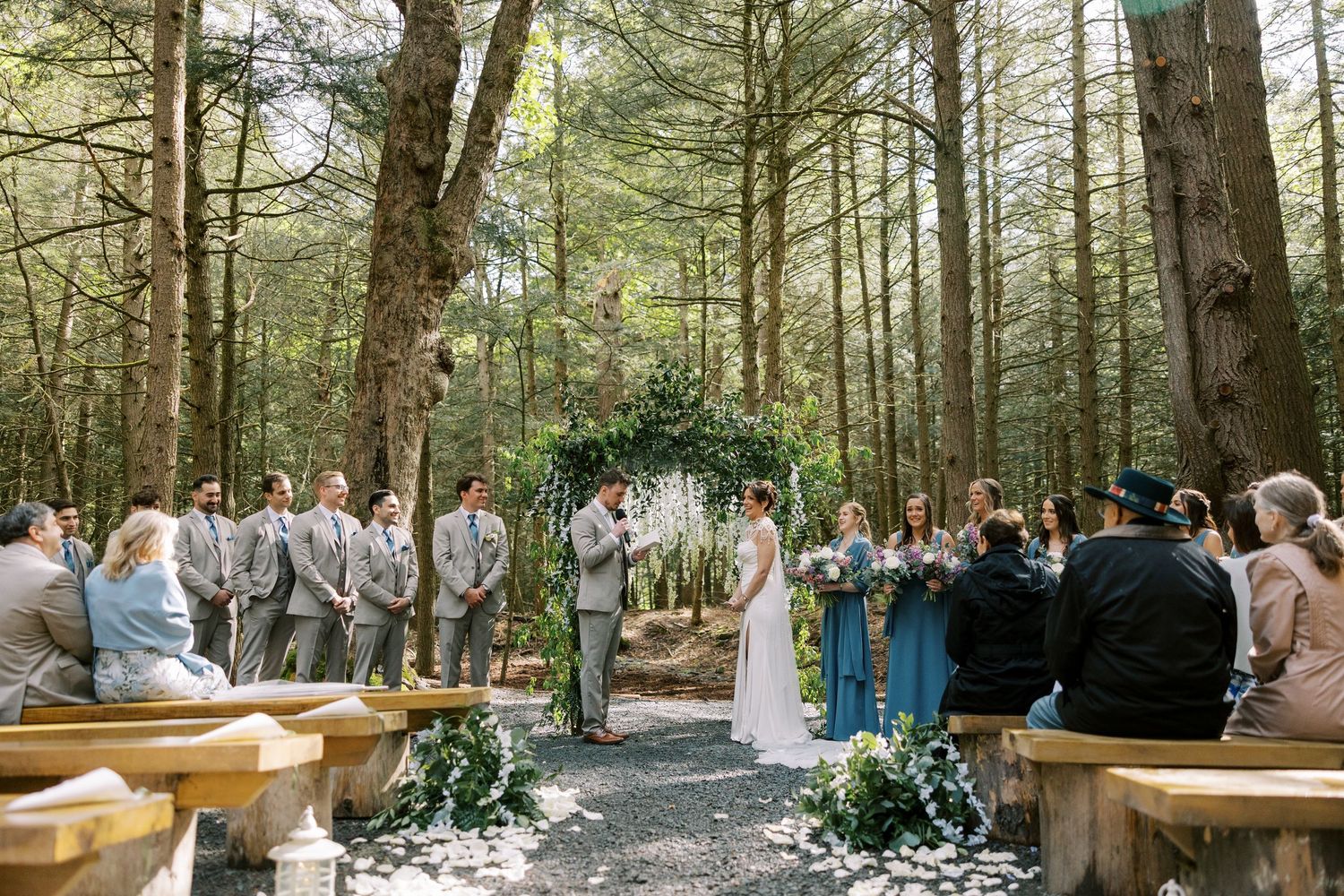 A woodland wedding ceremony with wooden benches and a floral arch set among tall pine trees in a forest clearing at Windham Manor.