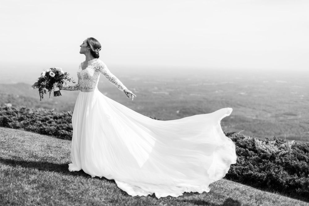 A flowing white wedding dress billows dramatically in the wind against a scenic mountain landscape at The Cliffs at Glassy Chapel.