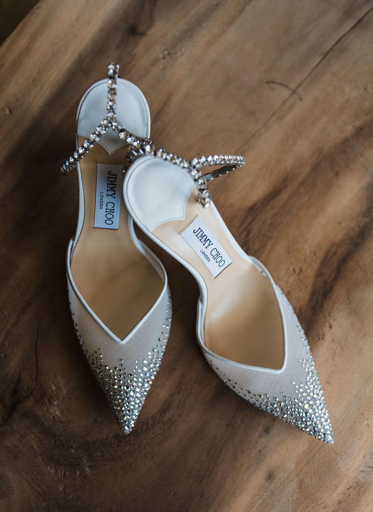 Elegant Jimmy Choo white bridal shoes with crystal embellishments on pointed toes displayed on wooden floor.