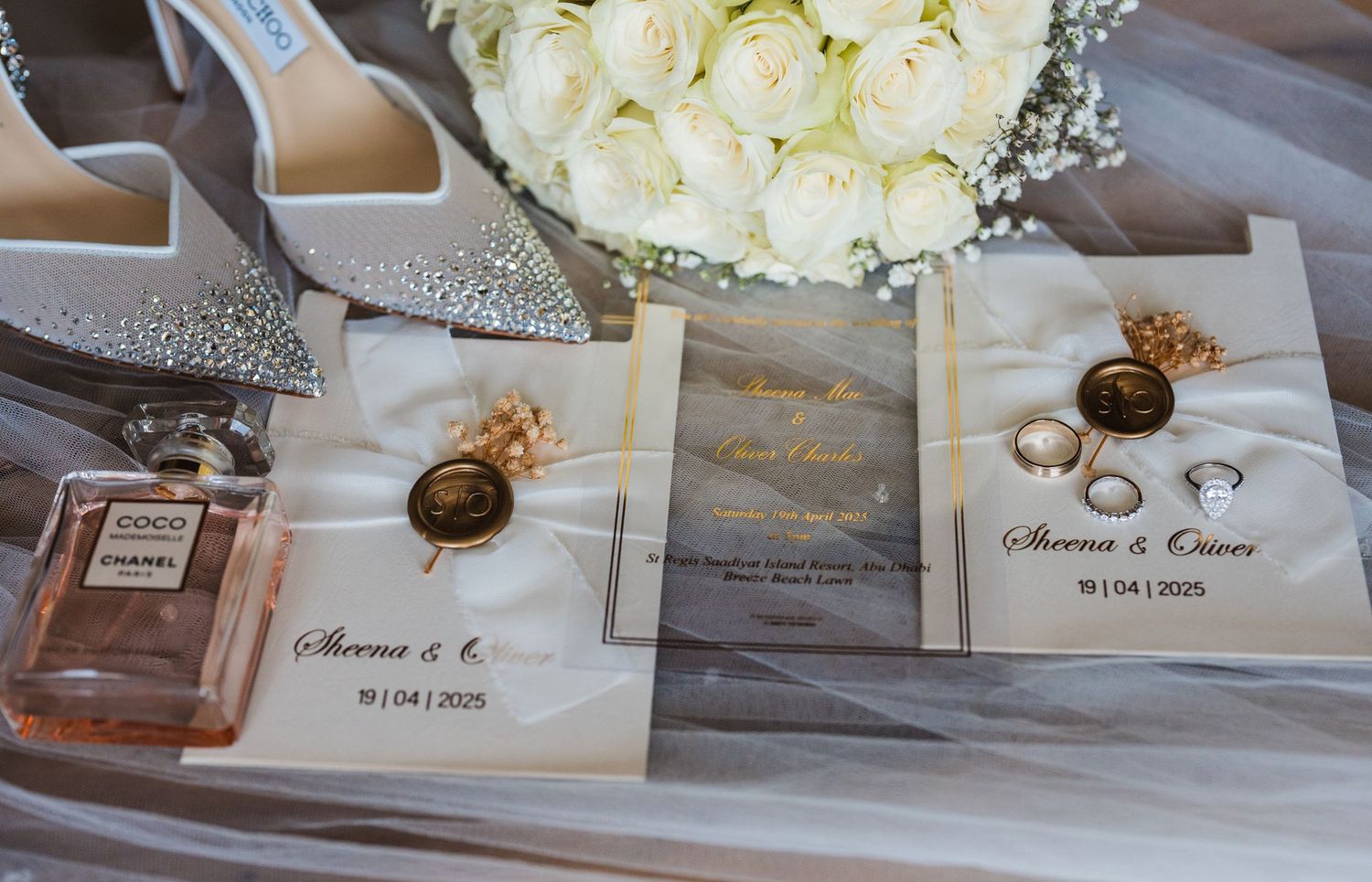 Wedding details including rings on invitations, perfume, silver shoes and white rose bouquet arranged on gray surface.