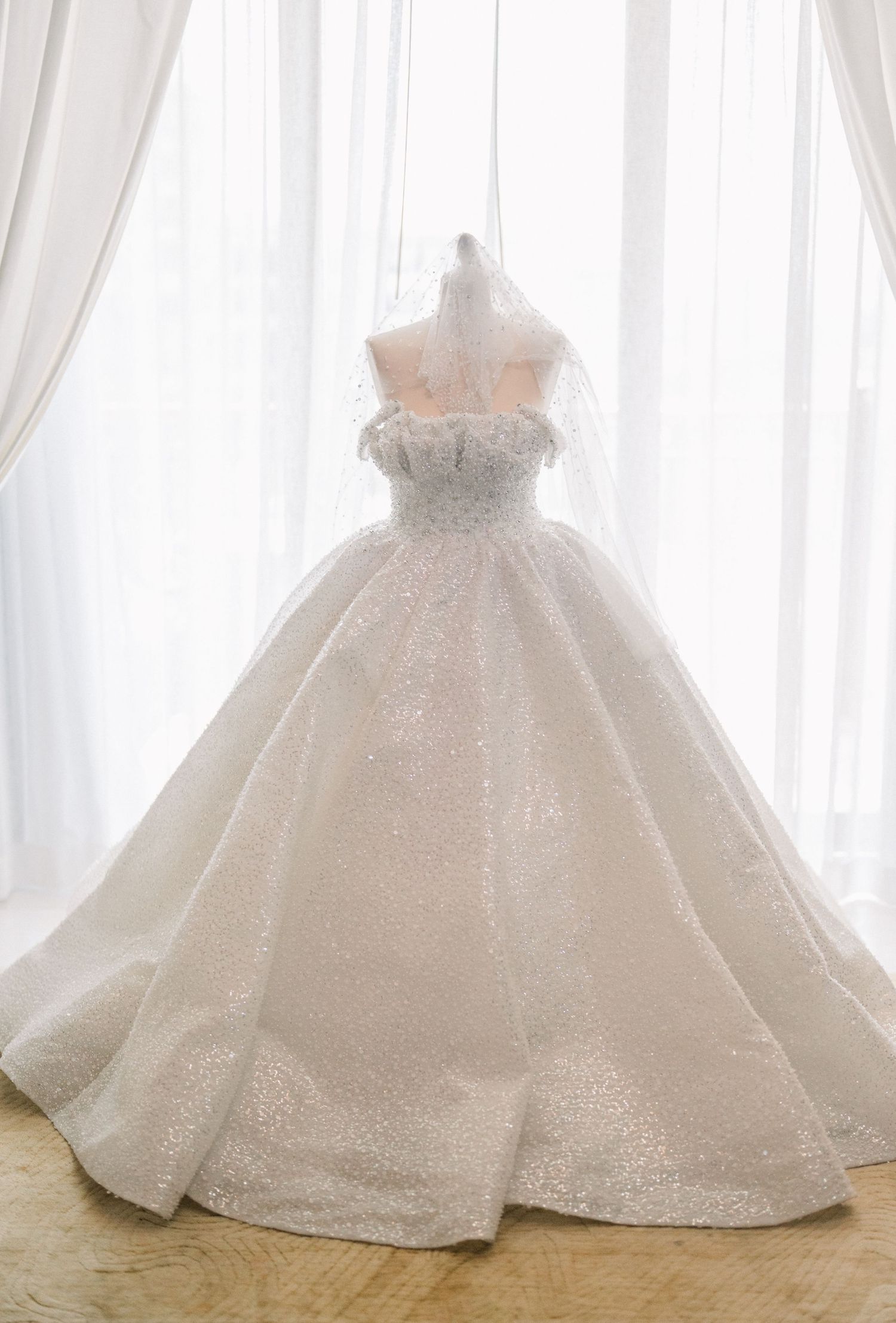 A luxurious white ball gown wedding dress with strapless bodice and full skirt displayed against sheer curtains.