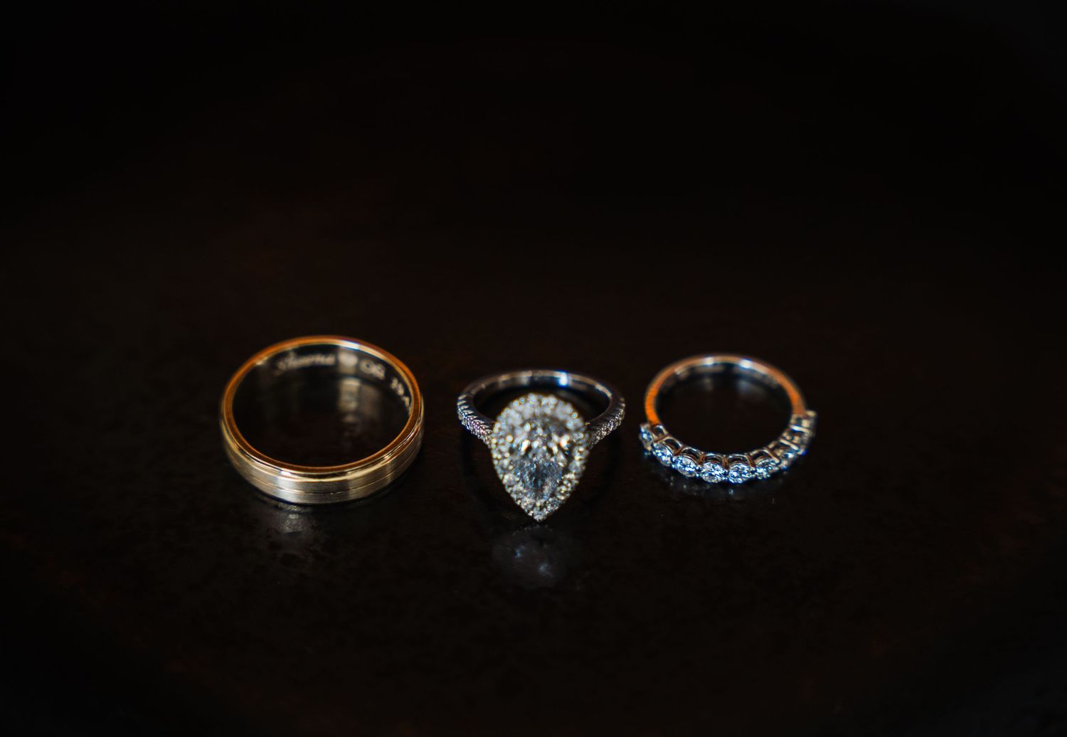 Three elegant wedding rings including a pear-shaped diamond engagement ring and blue gemstone band on dark background.