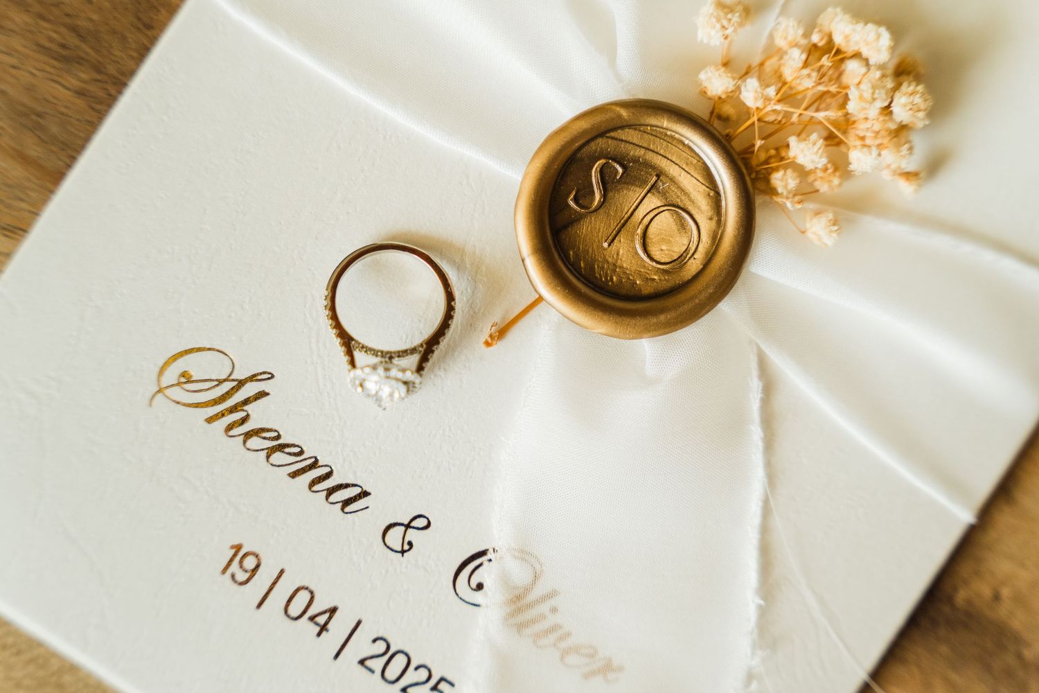 Wedding invitation with gold lettering, engagement ring, and wax seal on white fabric with delicate floral details.