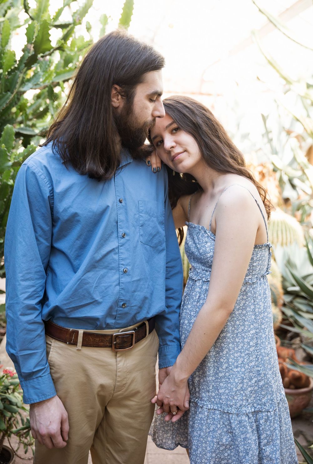 A romantic outdoor portrait of a couple in casual attire sharing an intimate moment among greenery and natural light.