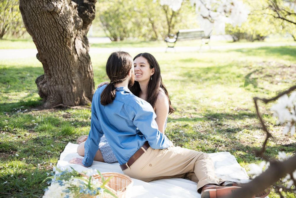 A couple enjoying a picnic date on a blanket under a large tree in a park setting.