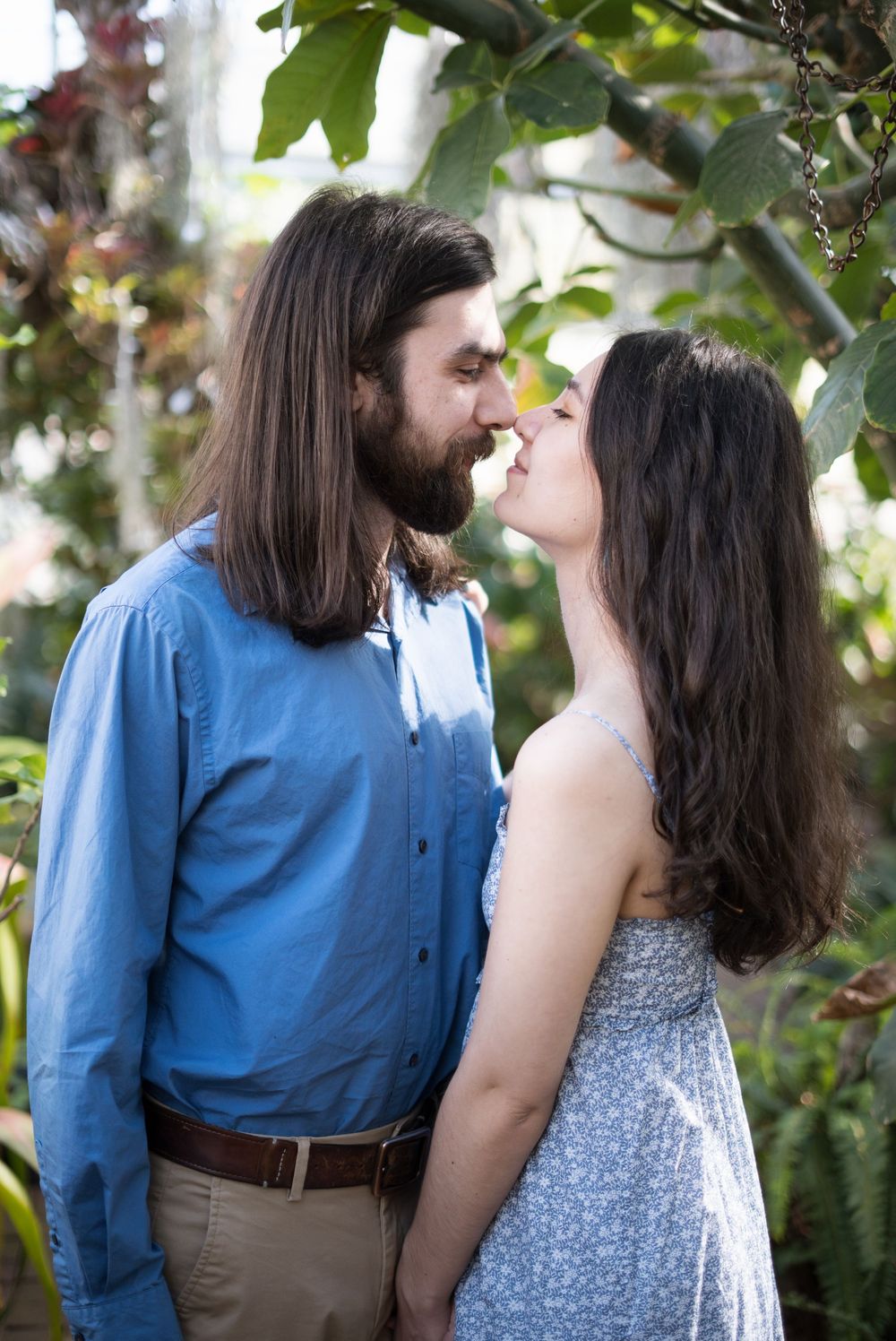A romantic garden portrait of a couple in casual attire gazing at each other among green foliage.