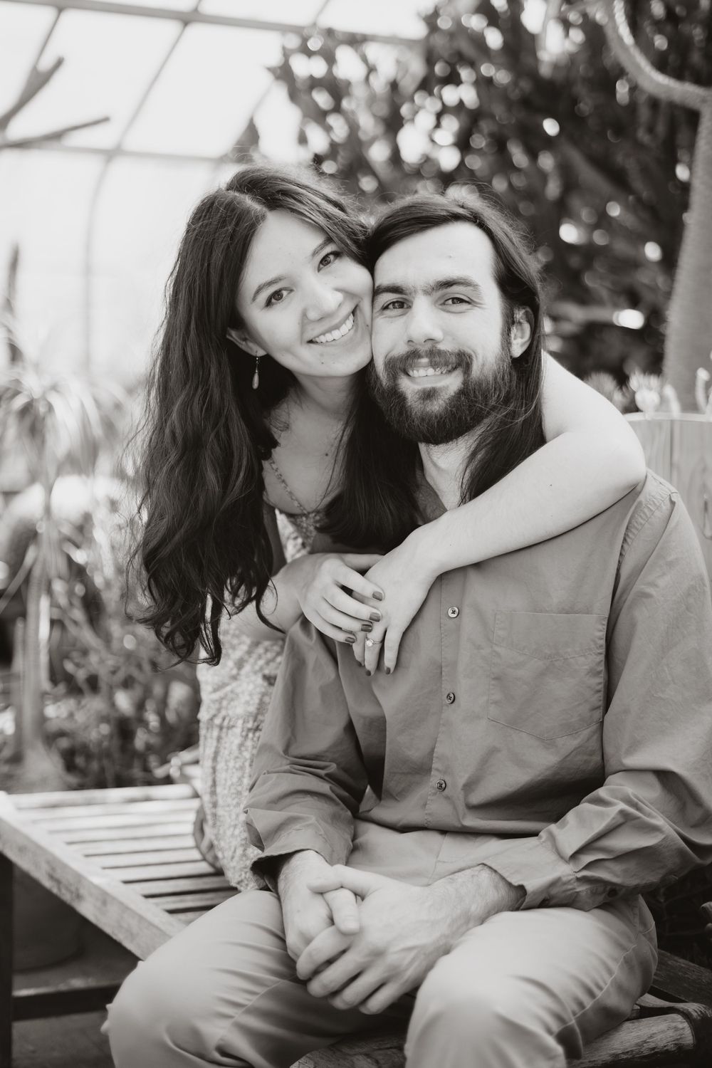 A black and white portrait of a couple sharing a warm embrace while sitting on a wooden bench outdoors.