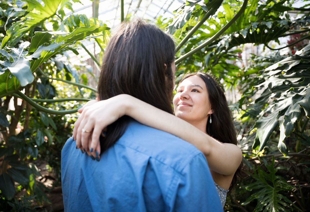 A couple embraces warmly in a lush greenhouse surrounded by tropical green plants and natural light.