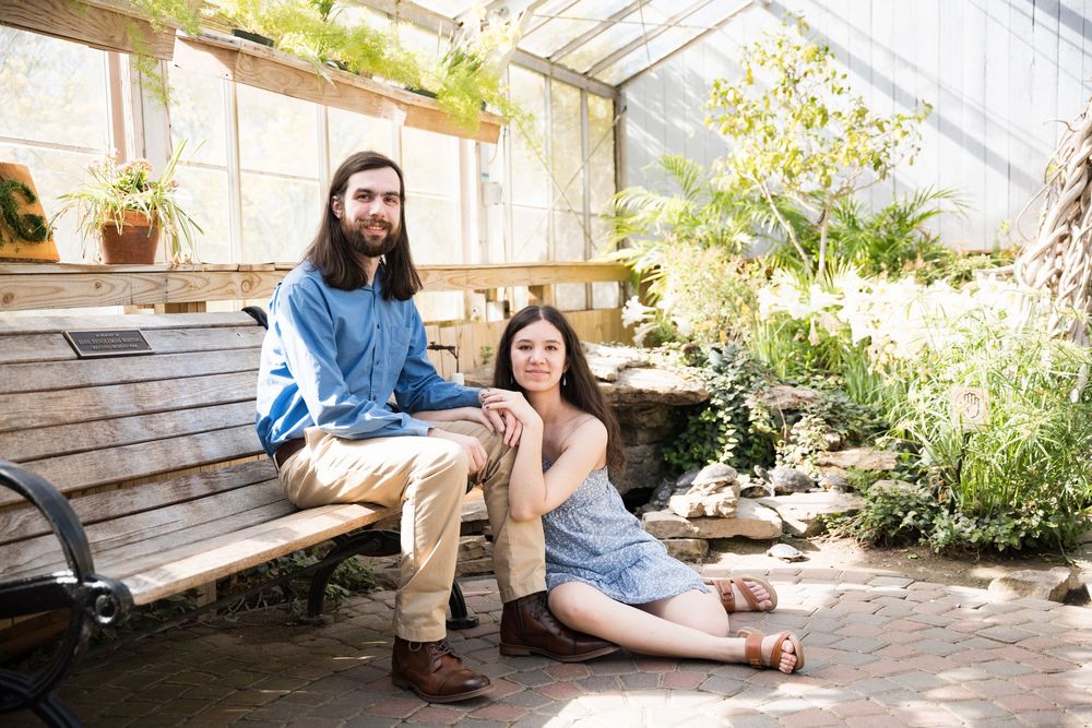 Two people sit together in a greenhouse surrounded by potted plants and natural lighting on a wooden bench.