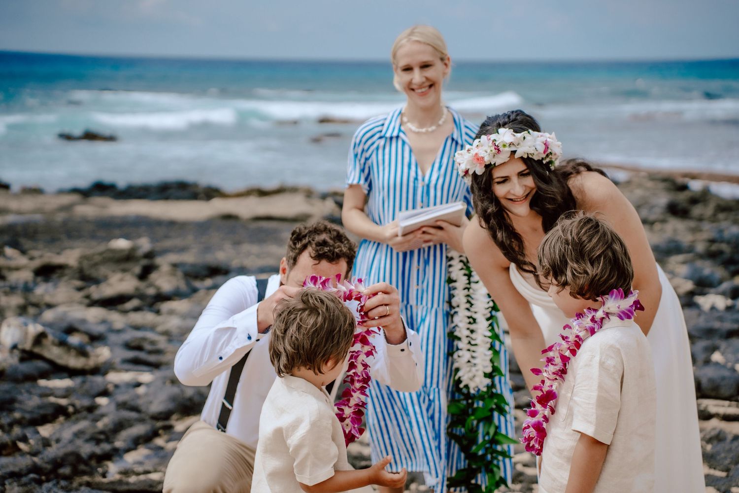 A group on a rocky Hawaiian beach wearing flower leis and traditional wedding attire gather together by the ocean.