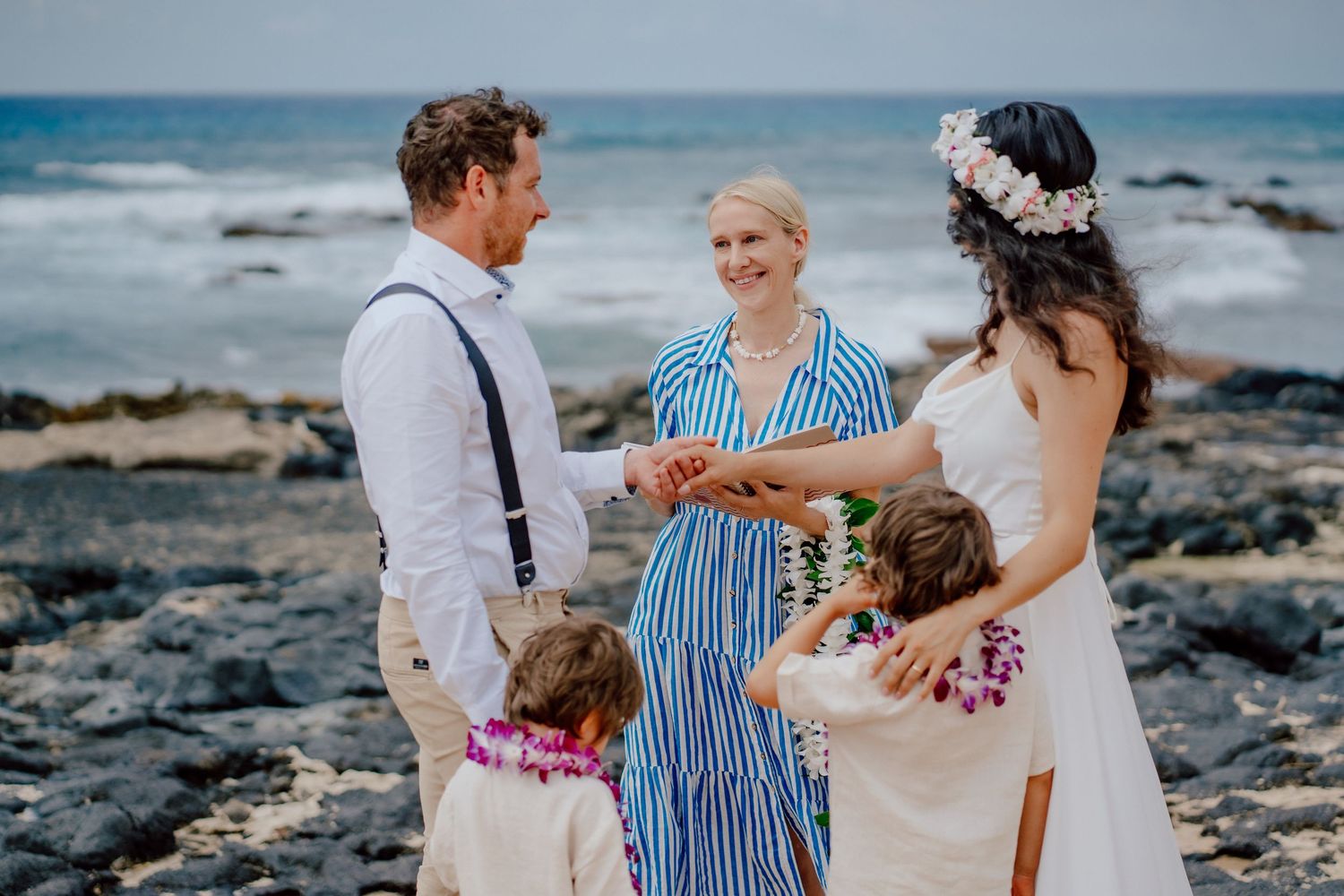 Intimate beach wedding ceremony takes place on rocky Hawaiian coastline with waves crashing in background.