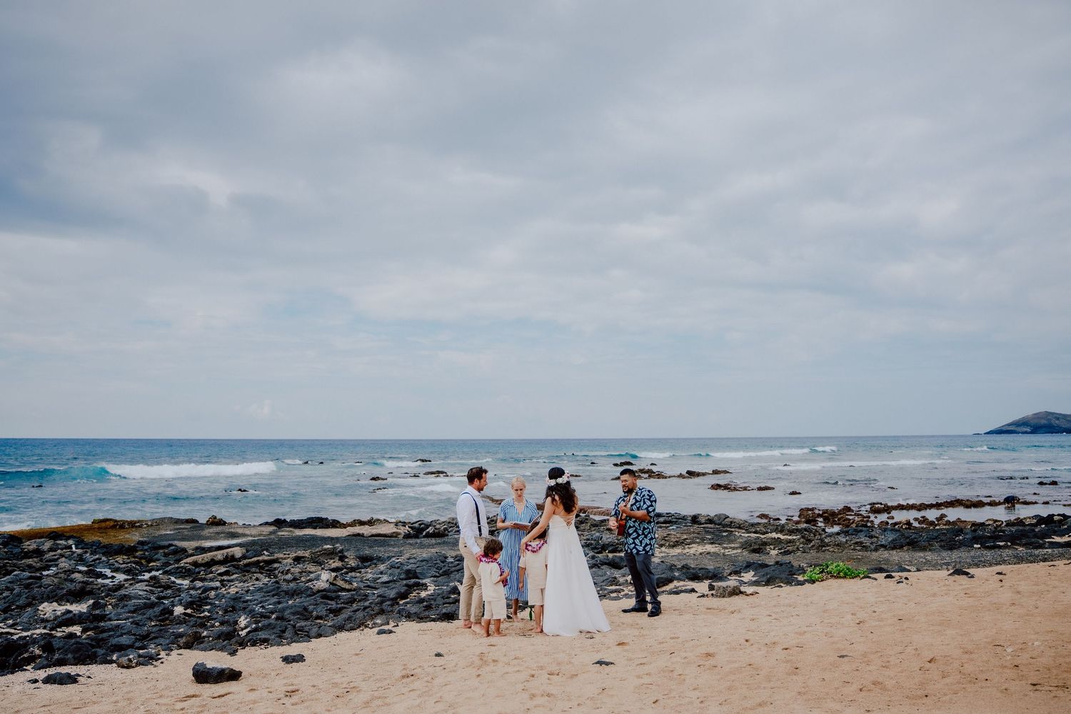 Small wedding ceremony takes place on a rocky beach shore in Hawaii with ocean waves and cloudy sky in background.
