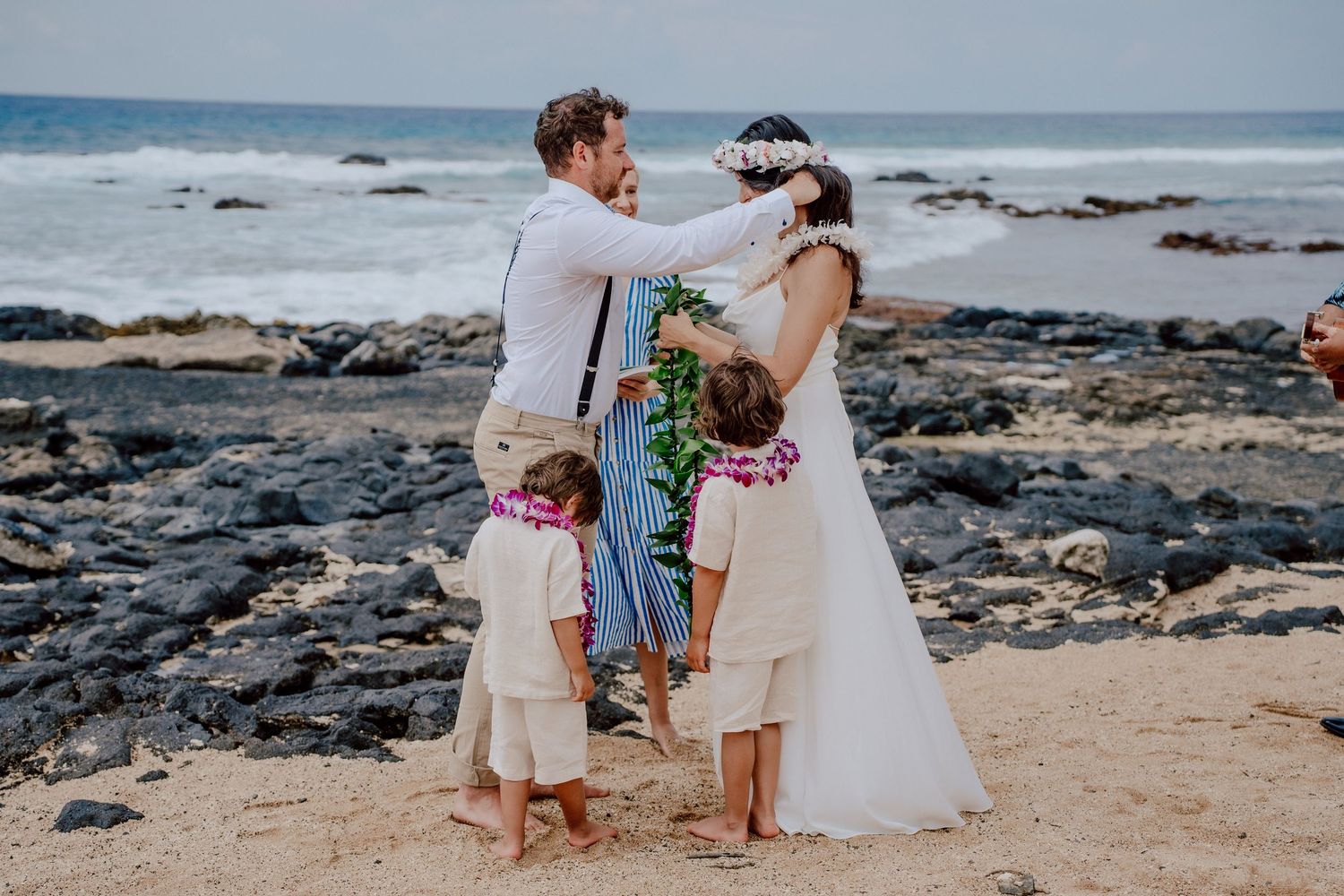 A beach wedding celebration with ohana on a rocky Hawaiian shoreline as waves crash against volcanic rocks in the background.