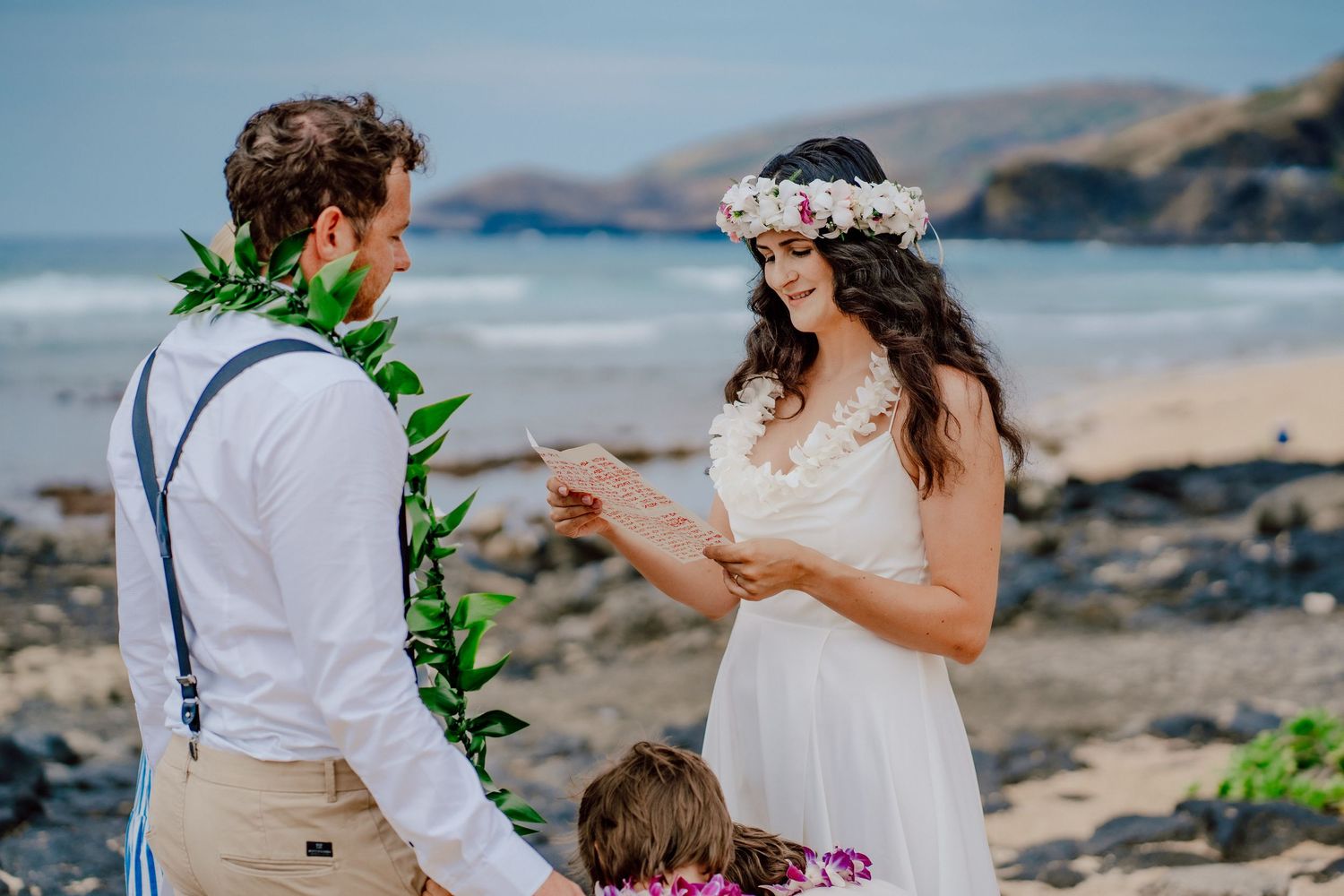 Couple exchanges vows during intimate beach wedding ceremony on rocky Hawaiian coastline with ocean and mountains behind them.