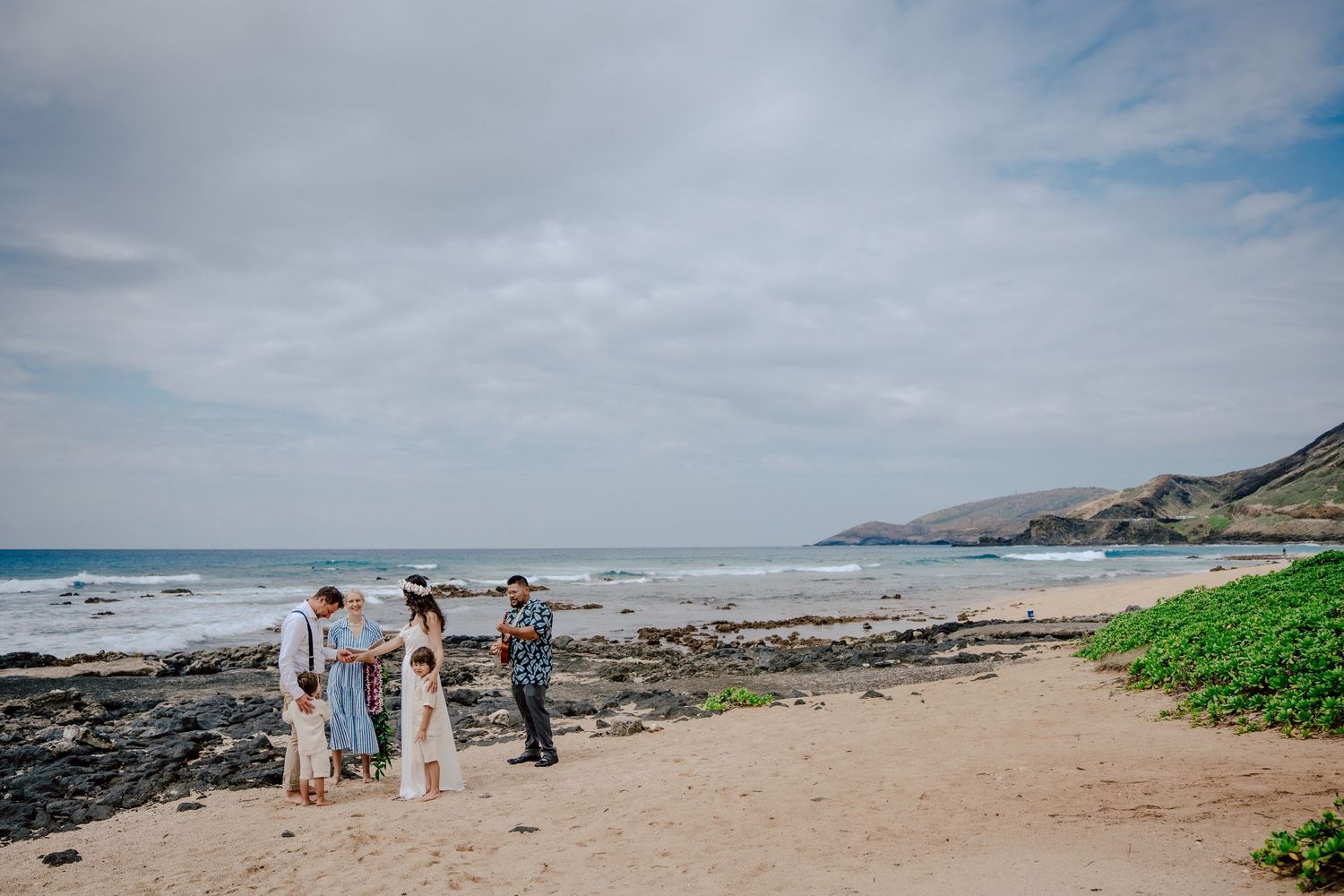 A group of people stand on a scenic rocky beach with turquoise ocean waters and mountains in the distance.