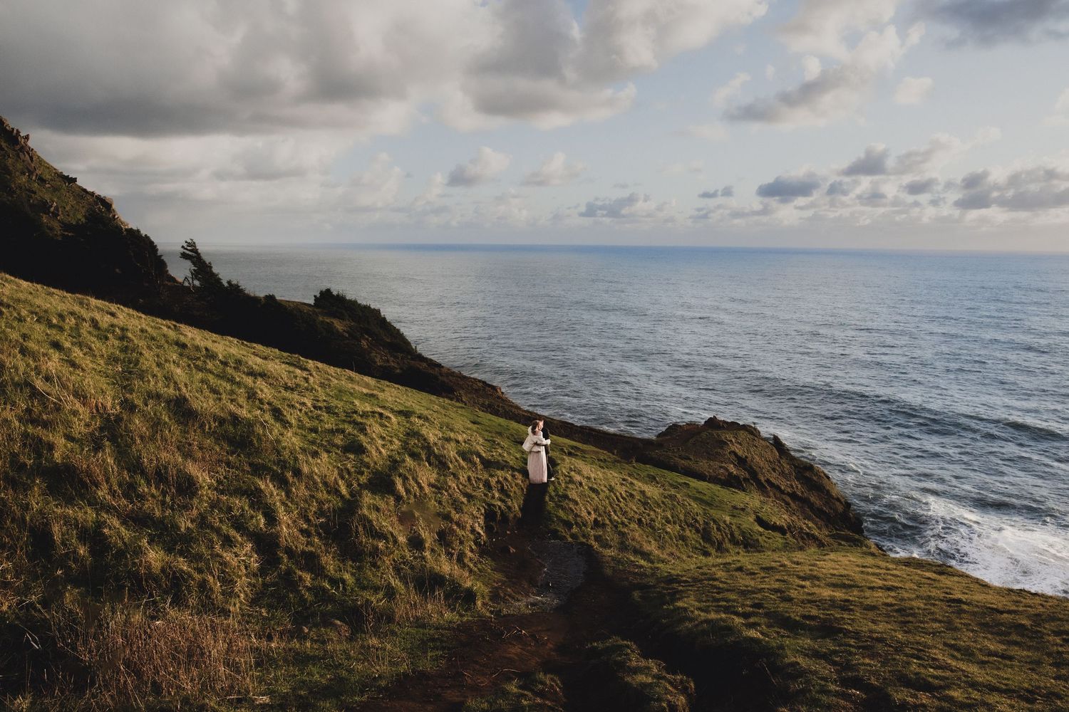 A lone figure walks along a grassy coastal cliff path overlooking the ocean under a dramatic cloudy sky.