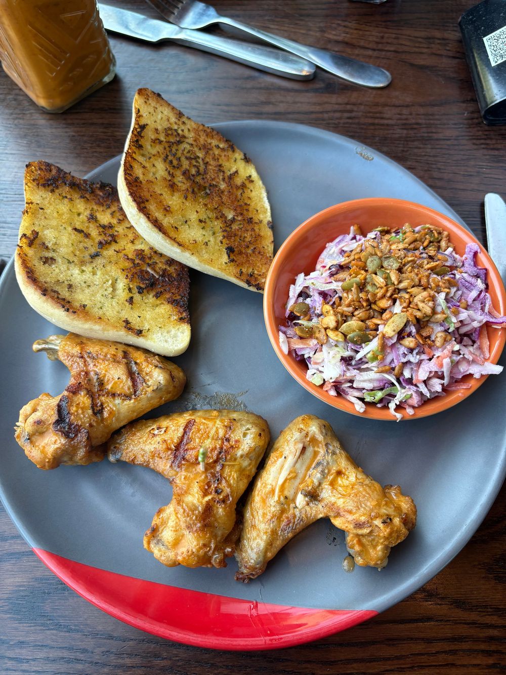 A plate of grilled chicken wings with garlic bread and a side of coleslaw in an orange bowl on a wooden table.