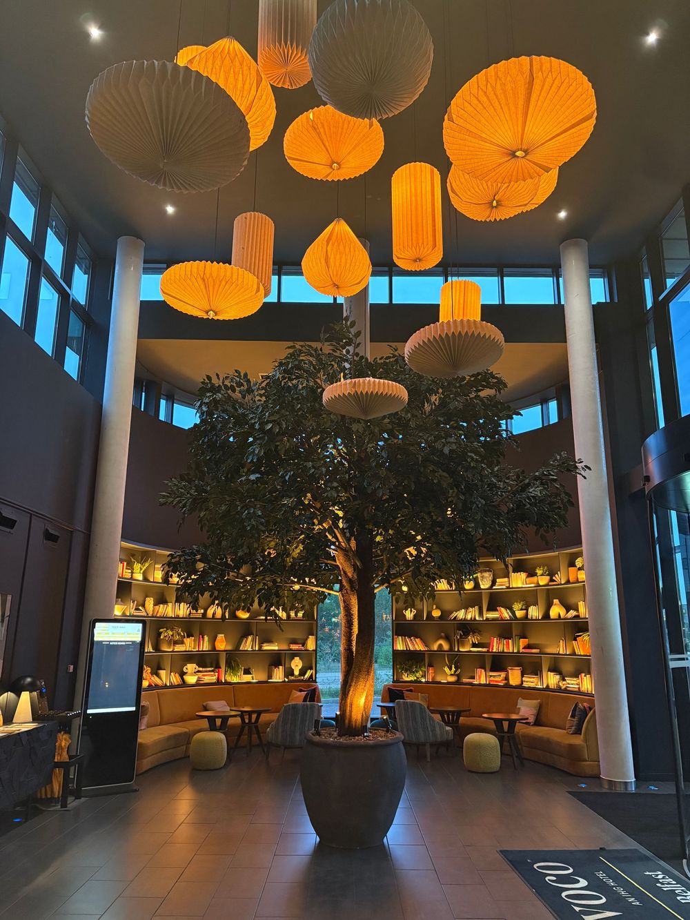 Modern restaurant interior with orange pendant lights, floor-to-ceiling windows, and large artificial tree centerpiece.